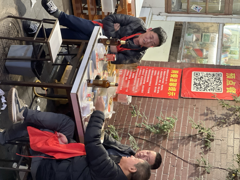 An outdoor dining scene in Yuzhong District, China, depicts three adult males seated around a rectangular table with a white top, positioned against a red brick wall. The wall is partially covered by climbing green vines and features two prominent red banners with white Chinese text; the upper banner displays a large QR code for "预点餐" (Pre-order meal), and the lower banner provides "用餐温馨提示" (Dining warm tips). To the left of the table, a lighted window display behind a wooden frame showcases various items on shelves.
On the left, a man wearing a black puffer jacket, eyeglasses, and a red apron is seated with his arms crossed, looking towards the right while speaking. Two brown glass beer bottles stand on the table in front of him. In the center-right, a man in a dark jacket is speaking, directing his gaze towards the third man. The third man, positioned on the far right, wears a black puffer jacket, dark pants, and grey-and-white sneakers, with a red apron draped across his lap. He is actively reaching into a food dish on the table.
The table is covered with multiple dishes of food, including bowls, plates, chopsticks, and crumpled napkins. Visible beverages include at least three brown glass beer bottles, an aluminum can, and a clear pitcher filled with liquid. The seating consists of white-topped stools. A grey plastic trash bin with a black liner is visible under the table, and a metal manhole cover is on the ground beneath the seating area. The men appear to be engaged in conversation and dining.