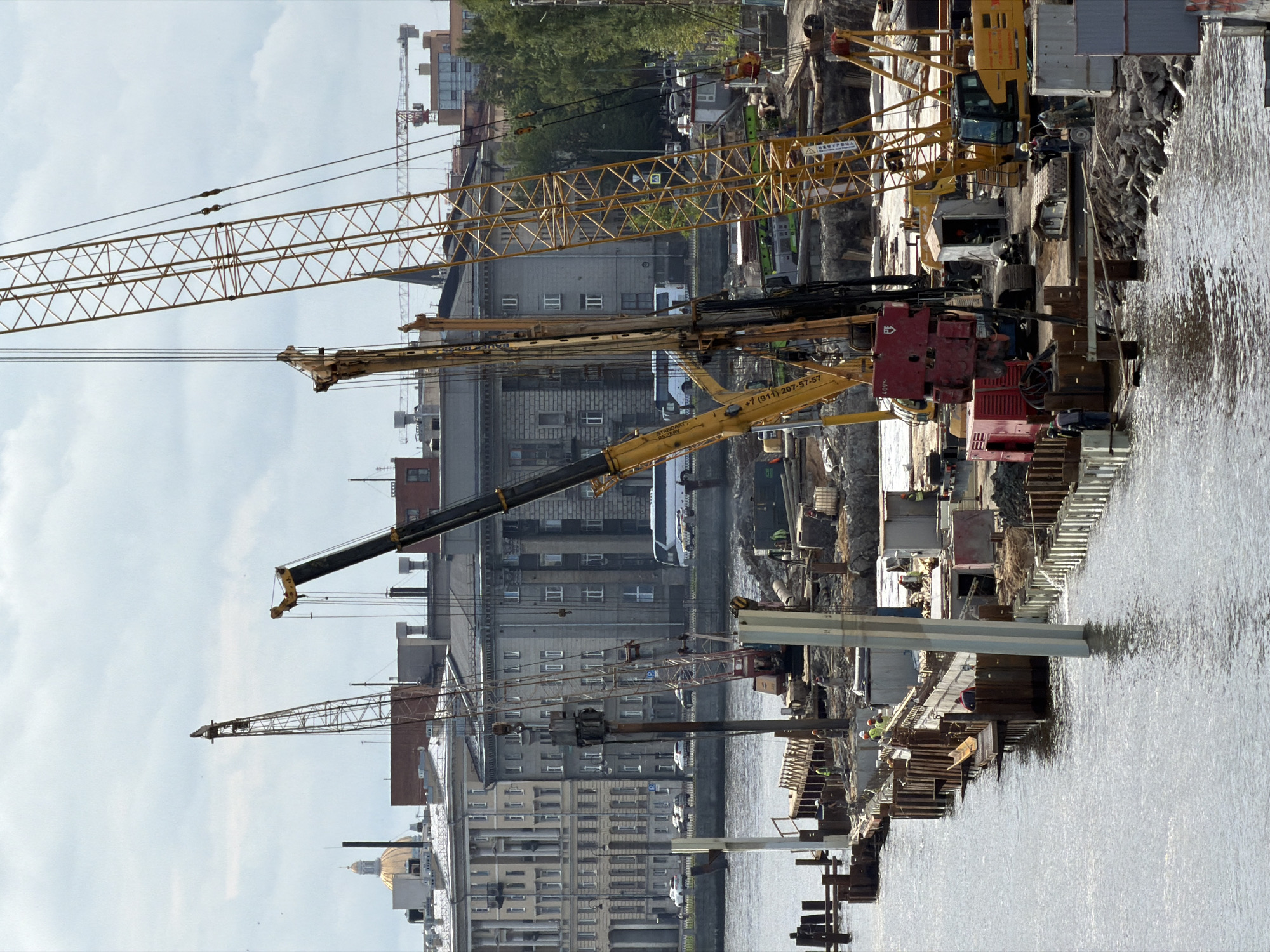 The image depicts an active construction site along the Neva River in Saint Petersburg, Russia, on a cloudy day. A large-scale infrastructure project is underway, evidenced by multiple construction cranes, drilling equipment, and piles of construction materials. The foreground features the Neva River with a section of the construction site extending into the water, marked by wooden pilings and a concrete barrier.
Several construction workers, dressed in safety vests and helmets, are visible on the site, engaged in various tasks. The middle ground reveals a complex arrangement of heavy machinery, including a bright yellow excavator, a red drilling rig, and two large tower cranes. In the background, grand, historical buildings with neoclassical architecture line the riverbank, characteristic of Saint Petersburg's cityscape. These buildings include structures with prominent columns and arched windows, and a distinct golden dome is visible in the distance. Cars and a bus are seen on a road running alongside the embankment, indicating the proximity of this construction zone to a public area. Trees and other vegetation frame the scene, suggesting the site is situated in a relatively green part of the city. The overall impression is one of ongoing urban development against a backdrop of historical architecture.