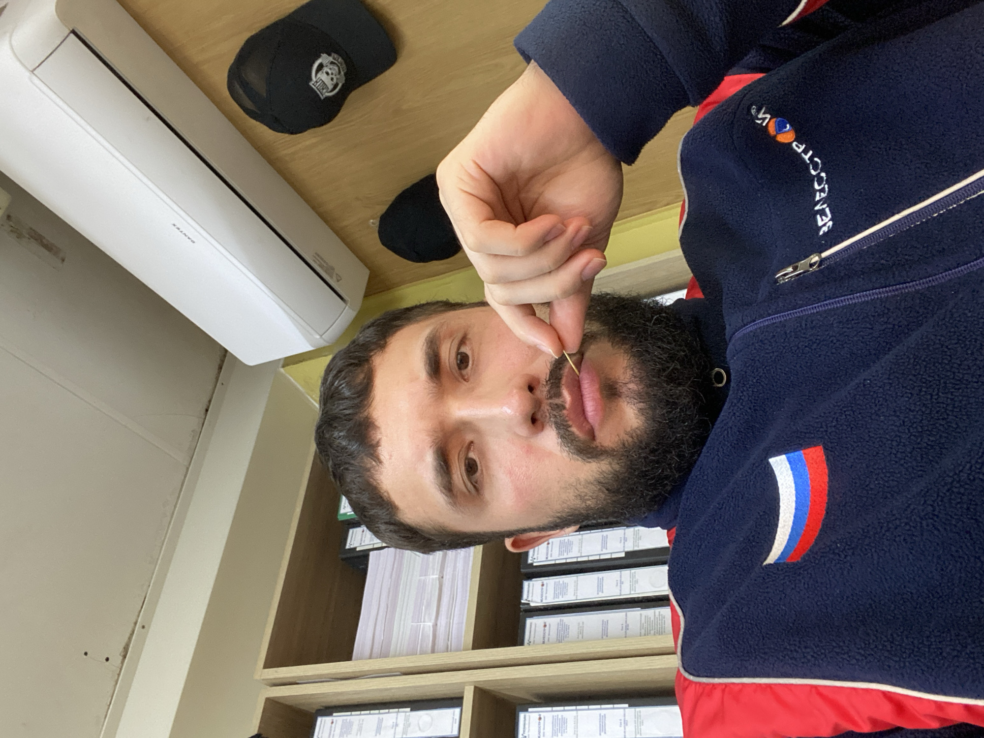 A man with a beard is sitting in an office in Luzhitsy, Russia. He is wearing a dark blue fleece jacket with red accents and a patch of the Russian flag on the left chest. He is holding a toothpick between his lips. Behind him, shelves are filled with binders. An air conditioner is mounted on the wall above him, and a black baseball cap with a skull logo is hanging on the wall to his right.