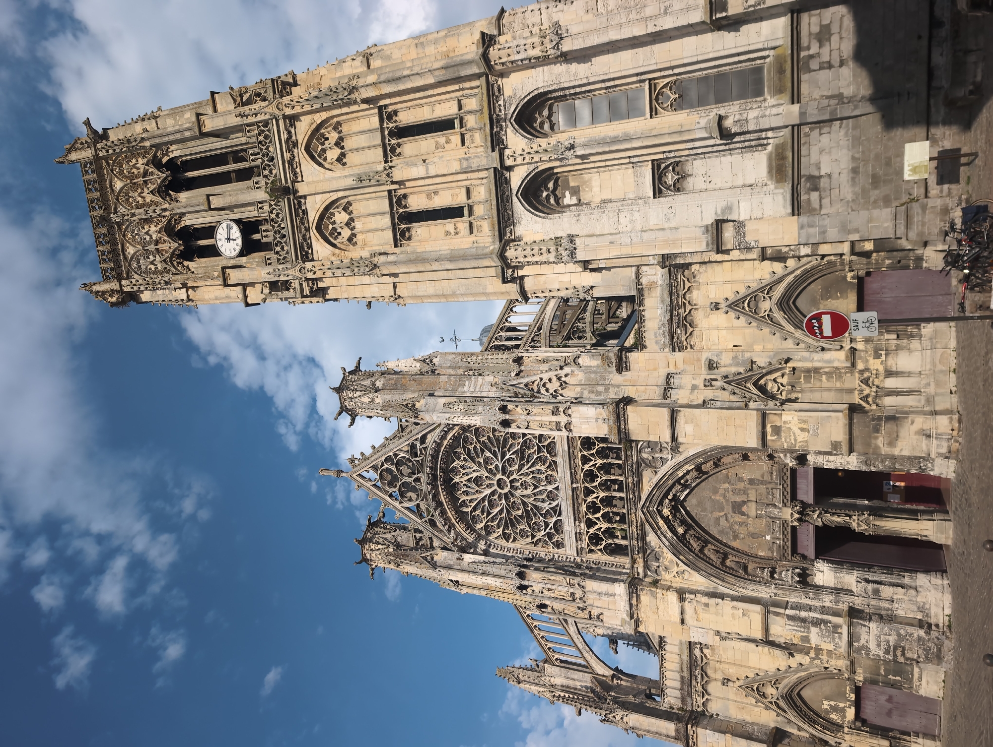 A wide-angle, low-angle exterior shot captures the imposing Gothic architecture of the Saint-Jacques Church in Dieppe, France. The church features a tall, ornate bell tower on the right, showcasing intricate stonework, arched windows, and a clock face. To the left of the tower, the main facade of the church is visible, dominated by a large, circular rose window above a grand arched entrance. The building is constructed of weathered light-colored stone. The sky above is a clear blue, dotted with white clouds. In the foreground, a cobblestone plaza is visible, with a sign indicating "SAUF CYCLISTES" (except cyclists) and a bicycle parked nearby. The scene is bathed in natural daylight, suggesting it was taken during the daytime.