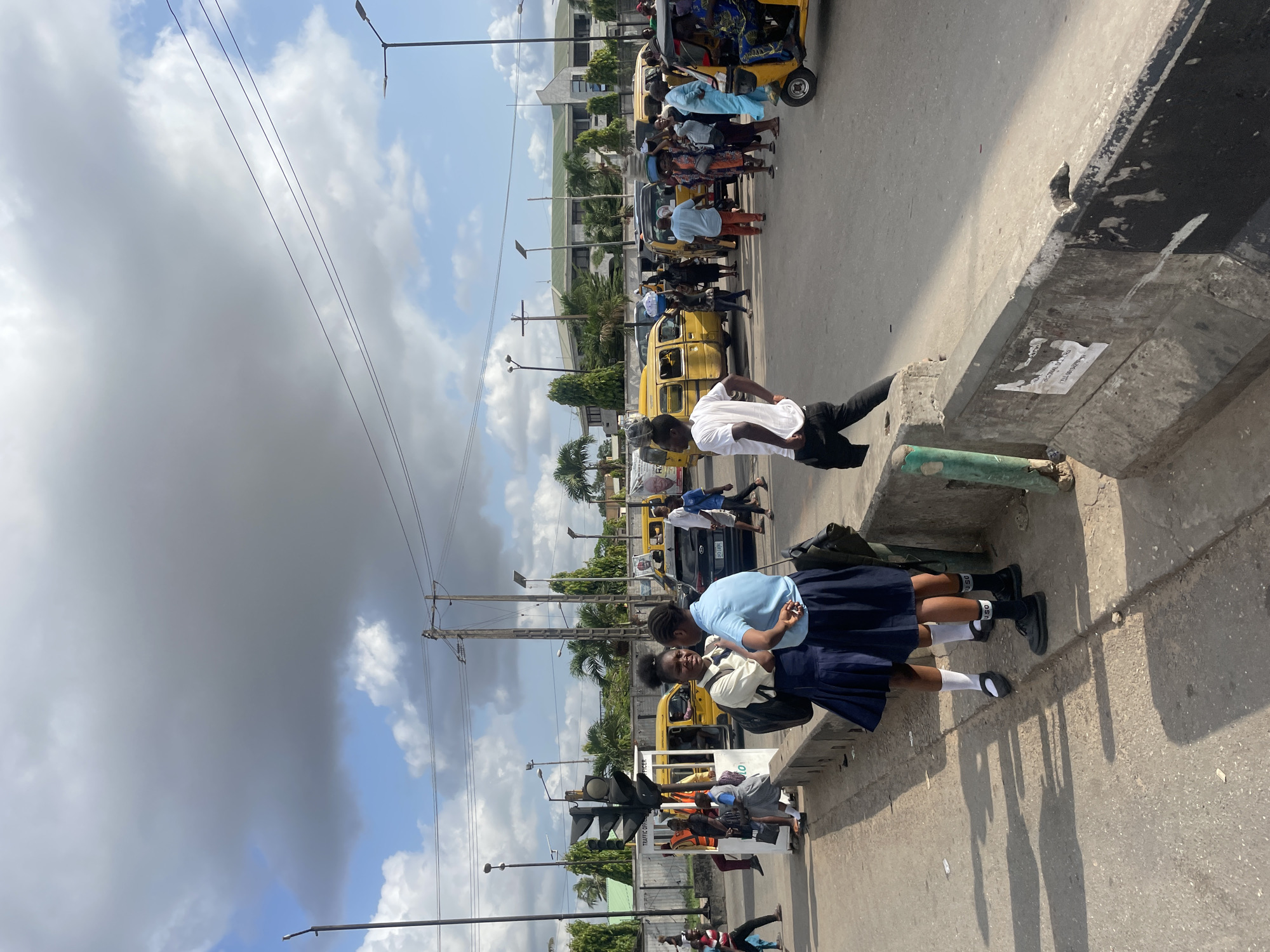Two schoolgirls in navy blue uniforms stand near a curb on a city street. The location is Ikeja, Nigeria based on uniform patches. Yellow "keke napep" auto rickshaws are in the background where people are gathered. Power lines and light poles are visible. The sky is partly cloudy.
