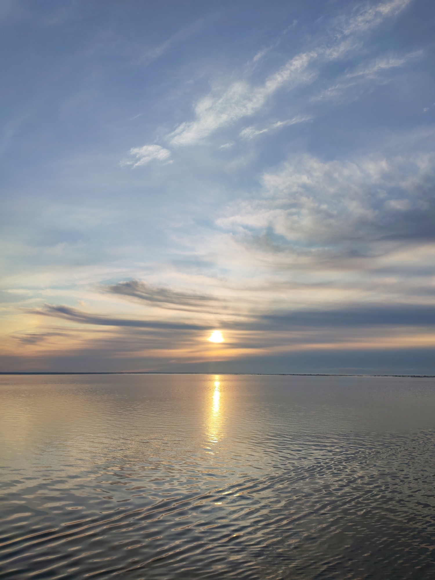The image depicts a serene seascape at sunset. The sky is a canvas of soft blues and grays, streaked with wispy clouds that catch the warm hues of the setting sun. The sun itself is a bright orb positioned low on the horizon, casting a shimmering golden path across the calm water. The water reflects the sky and the sun, creating a mirror-like effect with gentle ripples disturbing the surface. A faint, dark line on the horizon suggests the presence of distant land. The overall atmosphere is peaceful and tranquil. The location is specified as Veregonets, Russia, suggesting a body of water near the city during the golden hour. There are no people or specific objects identifiable in the scene.
