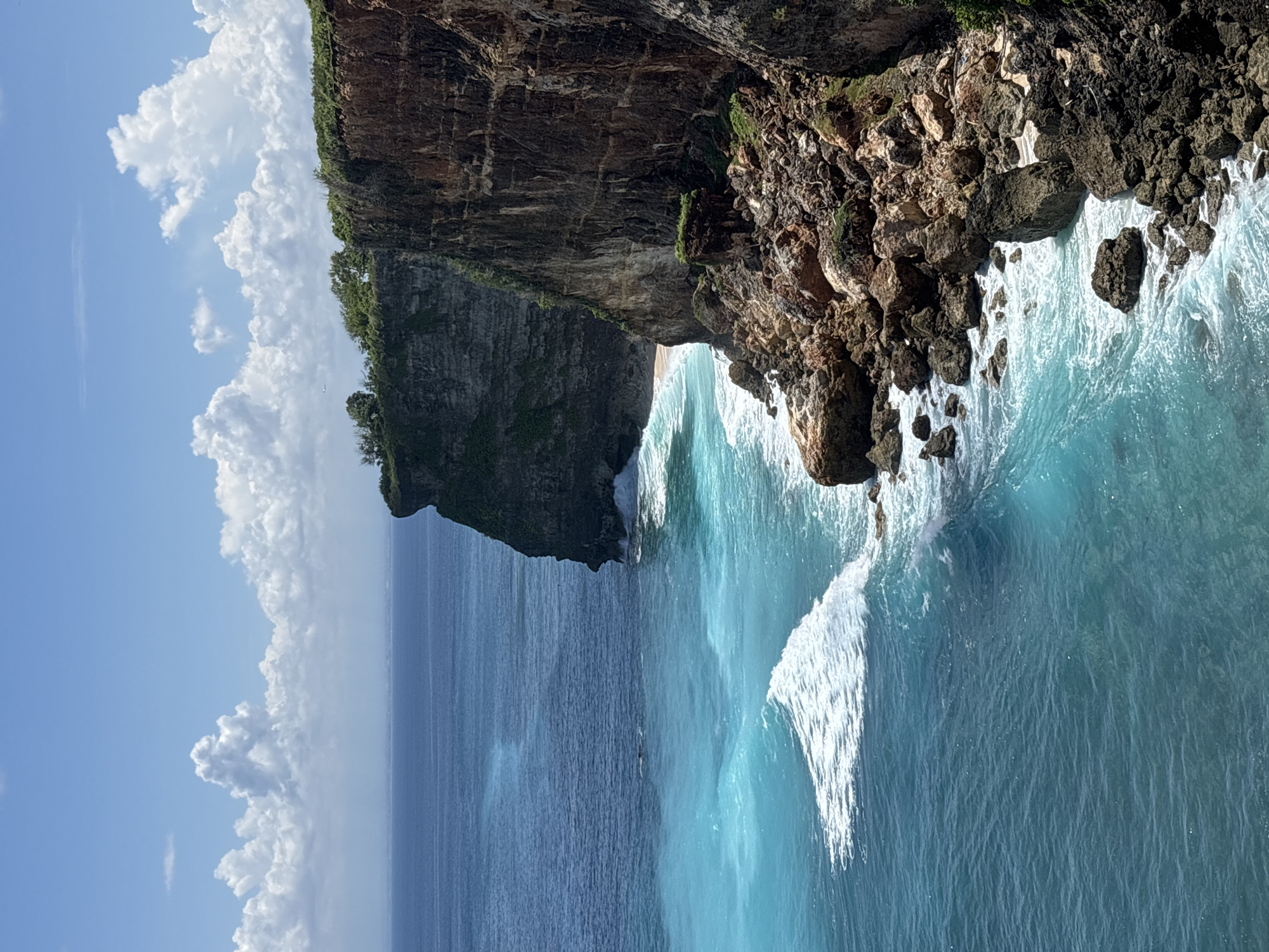 The image presents a coastal scene in Uluwatu, Indonesia, featuring a towering cliff face meeting the ocean. The cliff is comprised of layered rock formations, with vegetation clinging to its upper edge. The ocean is a vibrant turquoise, with white waves crashing against the base of the cliff and along the coastline. Cumulus clouds are visible in the blue sky above the scene. No people are visible.