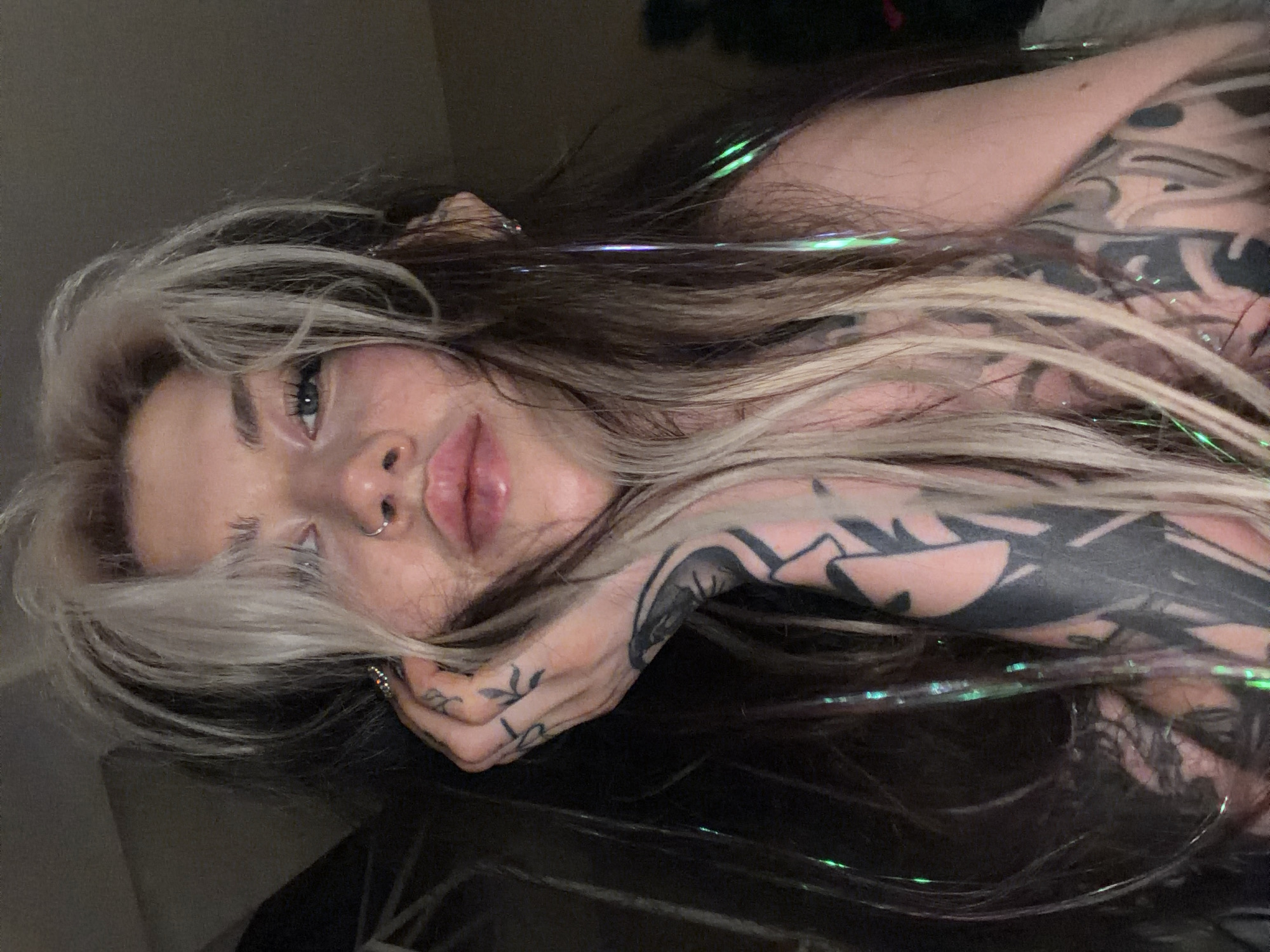 A woman with long, light-brown hair with blonde highlights poses for a picture. She has a nose ring and a tattoo on her hand. She is lying on a dark surface. She is not located in Budva, Montenegro.