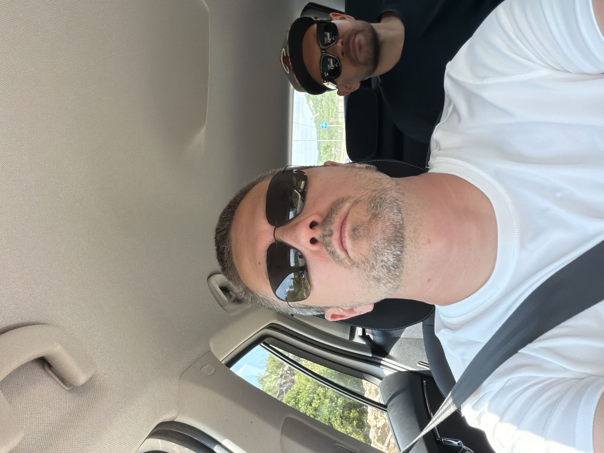 Two men are inside a car in Đuraševići, Montenegro. The man in the foreground is driving, wearing a white t-shirt and sunglasses. He has short gray hair and a goatee. The man in the background is in the passenger seat, wearing a black t-shirt and a baseball cap with sunglasses. The car's interior is visible, with the ceiling and part of the dashboard. Outside the car, through the windows, glimpses of a sunny day with greenery and rocky terrain can be seen.