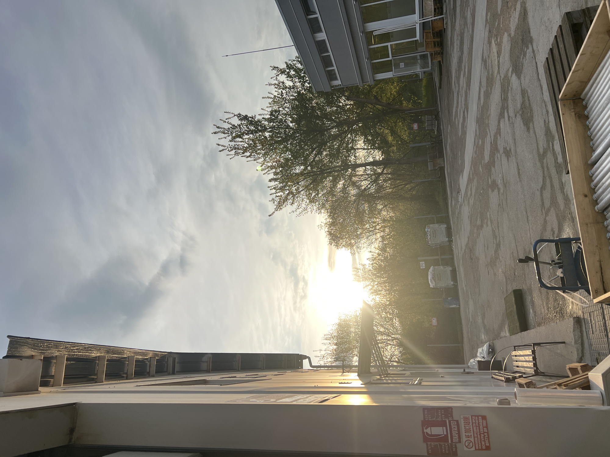 This image, captured in Sevlievo, Bulgaria, portrays an outdoor industrial or commercial yard during what appears to be late afternoon or early morning, marked by bright, low-angle sunlight. The sun, partially obscured by clouds and trees, creates a prominent lens flare, illuminating the scene with a warm glow.
On the left side of the frame, a white building or structure is visible. Affixed to its surface is a red and white warning sticker with text in Bulgarian. The legible parts of the text include: "ПОЖАРОГАСИТЕЛ" (Fire extinguisher), "НЕ ПОСТАВЯЙ! НИКАКВИ ПРЕДМЕТИ ПРЕД ПОЖАРОТЕХНИЧЕСКИТЕ СРЕДСТВА ОСИГУРИ СВОБОДЕН ДОСТЪП ДО ТЯХ!" (Do not place! No objects in front of the fire protection equipment. Ensure free access to them!). Other fragments like "КАТЕТО" and "ГРУПА" are also present.
Towards the right, a modern-style building with multiple windows stands, partly concealed by a cluster of trees with green foliage. A slender pole, possibly an antenna or lightning rod, extends from the roof of this building. The ground is a paved or concrete surface, showing signs of wear and various markings.
Numerous objects are distributed throughout the yard. In the immediate right foreground, a wooden crate contains a stack of long, cylindrical items, resembling fluorescent light tubes. Further into the scene, a blue and black hand truck or cart is parked. In the background, several large white bags or containers (likely bulk bags) are visible, along with stacked wooden pallets and construction materials, including what appear to be window frames or panels. The setting is absent of people, suggesting a functional, working environment.