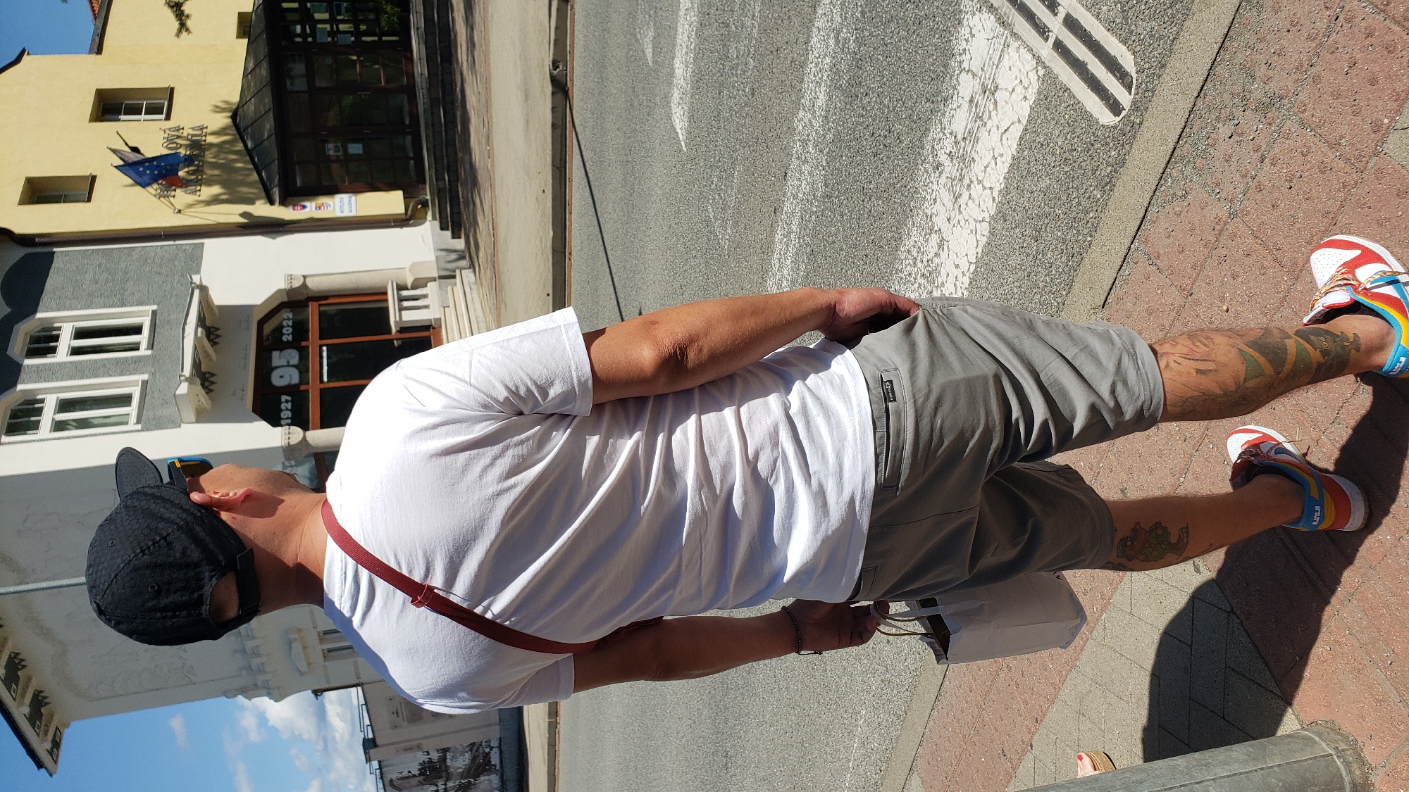 A man stands on a sidewalk in front of a building with a sign that reads "1927 95 2022." He is wearing a white t-shirt, gray shorts, and a black baseball cap. A red strap is visible across his chest, and he is carrying a white paper bag in his left hand. The building appears to be a historical structure, possibly an academy, with a yellow facade on the right side. There are flags on the building, including the European Union flag. The man has tattoos on his legs, and he is wearing colorful sneakers. The background shows a street with a pedestrian crossing. The scene is set in the city of Prešov, Slovakia.