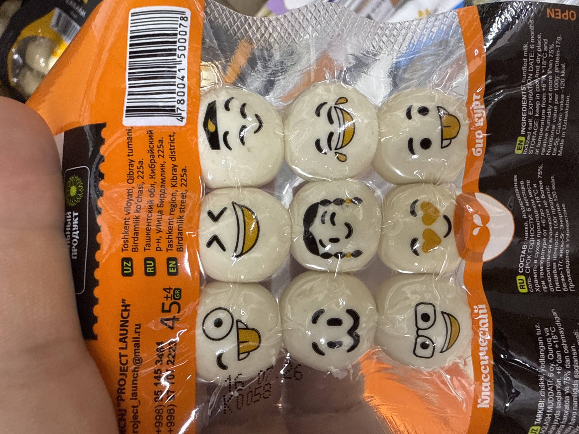 The image shows a close-up view of a bag of candies with emoji faces. The bag is orange and black with text in Russian, Uzbek, and English. Some of the text indicates that the product is from Tashkent, Uzbekistan. The candies are round and white with different emoji designs, such as winking faces, laughing faces, and faces with glasses. The bag also features a barcode and expiration date information. The candies are arranged in three rows of three. The bag appears to be sealed and has a glossy finish. The background is blurred and suggests an indoor environment, possibly a store shelf.