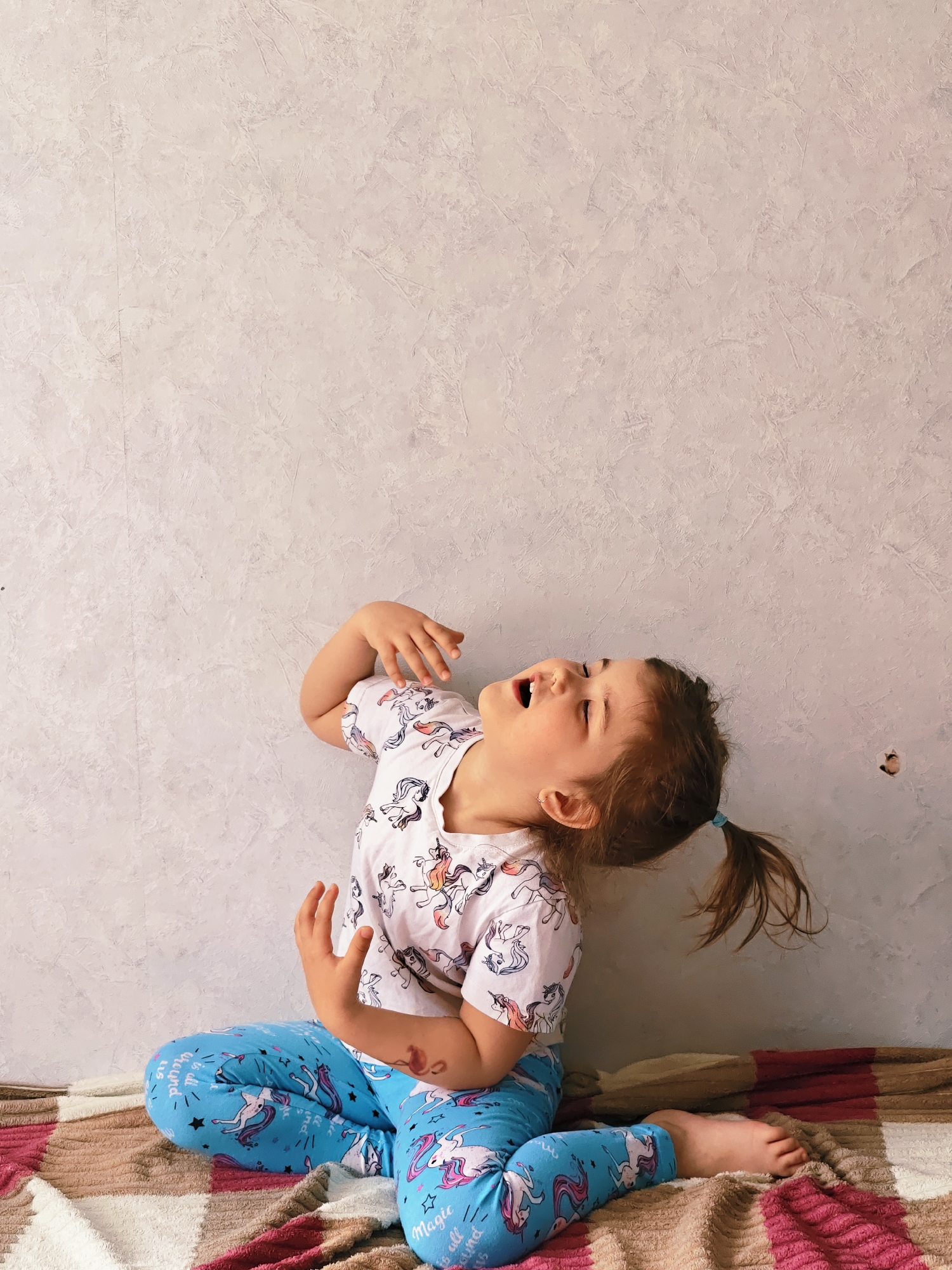 A young girl with brown hair in a ponytail is sitting cross-legged on a blanket with a plaid pattern in shades of white, tan, and red. She is wearing a white T-shirt with a unicorn pattern and blue pajama pants also decorated with unicorns, stars, and text. The girl is tilted back, looking upwards with her mouth open and both hands raised. She is positioned in front of a textured, light gray wall. The background appears to be an indoor setting. Based on the given information, the location is in the city of Chaikovsky, Russia.
