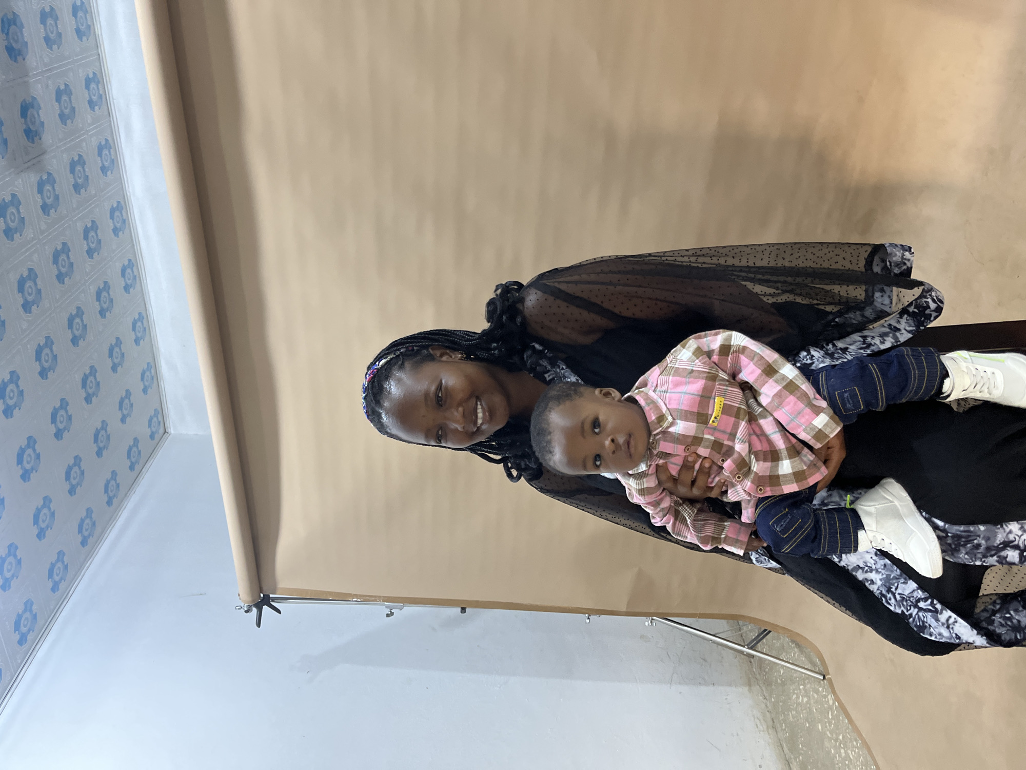 A Black woman, with dark braided hair adorned with a colorful headband, smiles at the camera. She is wearing a black sheer dress with polka dots over a patterned inner garment. She is holding a young Black child, who is looking directly at the camera. The child is wearing a pink and white checkered shirt and blue jeans with white sneakers. They are in front of a seamless tan backdrop, with a white wall and a patterned ceiling visible in the background. A photographic lighting stand is positioned to the left of the frame. The setting appears to be a studio. This image was taken in Gura Top, Nigeria.