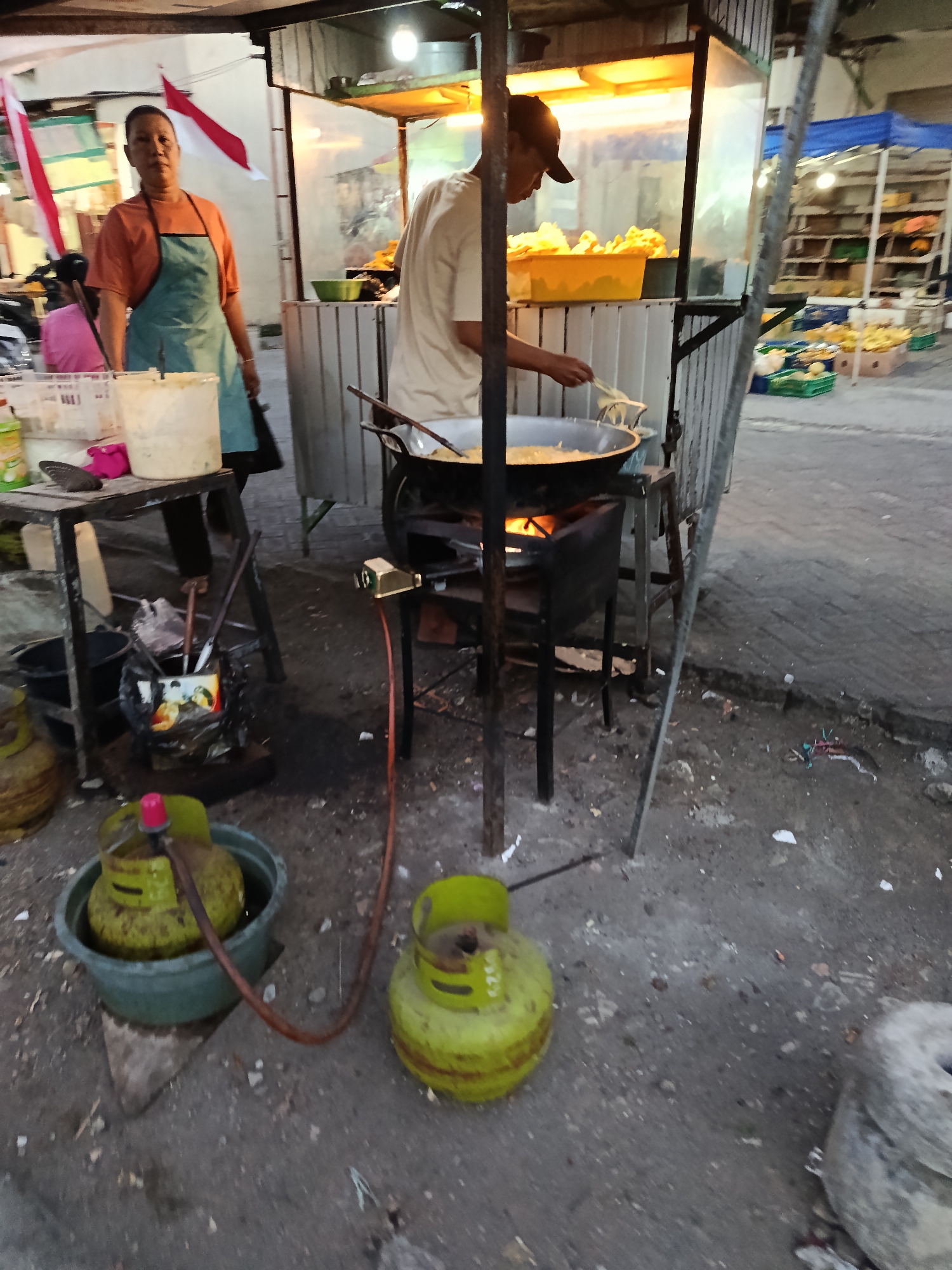 In Keboansikep, Indonesia, a vendor prepares food at an outdoor stall. A woman in an orange t-shirt and teal apron stands behind a table with a white bucket. A man wearing a light-colored t-shirt and a dark baseball cap is cooking in a large pan over a gas stove. The stall is equipped with a wok, gas tanks, and various containers. In the background, shelves display produce, and a blue canopy provides shade. The ground is paved with concrete and scattered with debris.