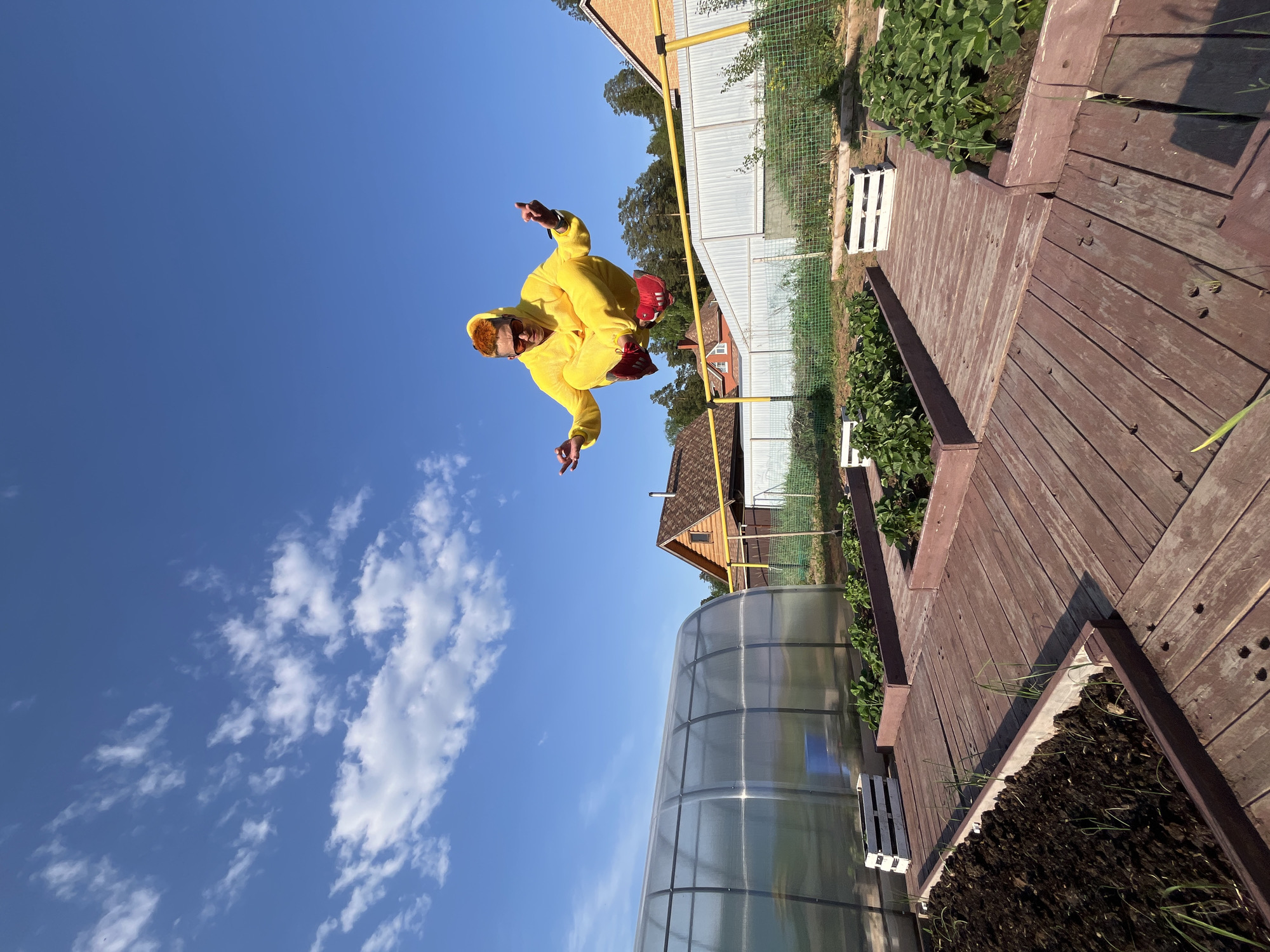 The image shows a person in a yellow jumpsuit and red sneakers seemingly jumping or posing in mid-air against a bright blue sky with scattered clouds. The location is likely a backyard in a rural area, possibly Chaikovsky, Russia, as indicated by the presence of typical Russian countryside elements.
In the background, there are several buildings with pitched roofs, a greenhouse, a wooden walkway with planters, and a chain-link fence. A yellow gas pipe is visible along the roof of one of the buildings. The ground appears to be mostly covered in grass and garden foliage.