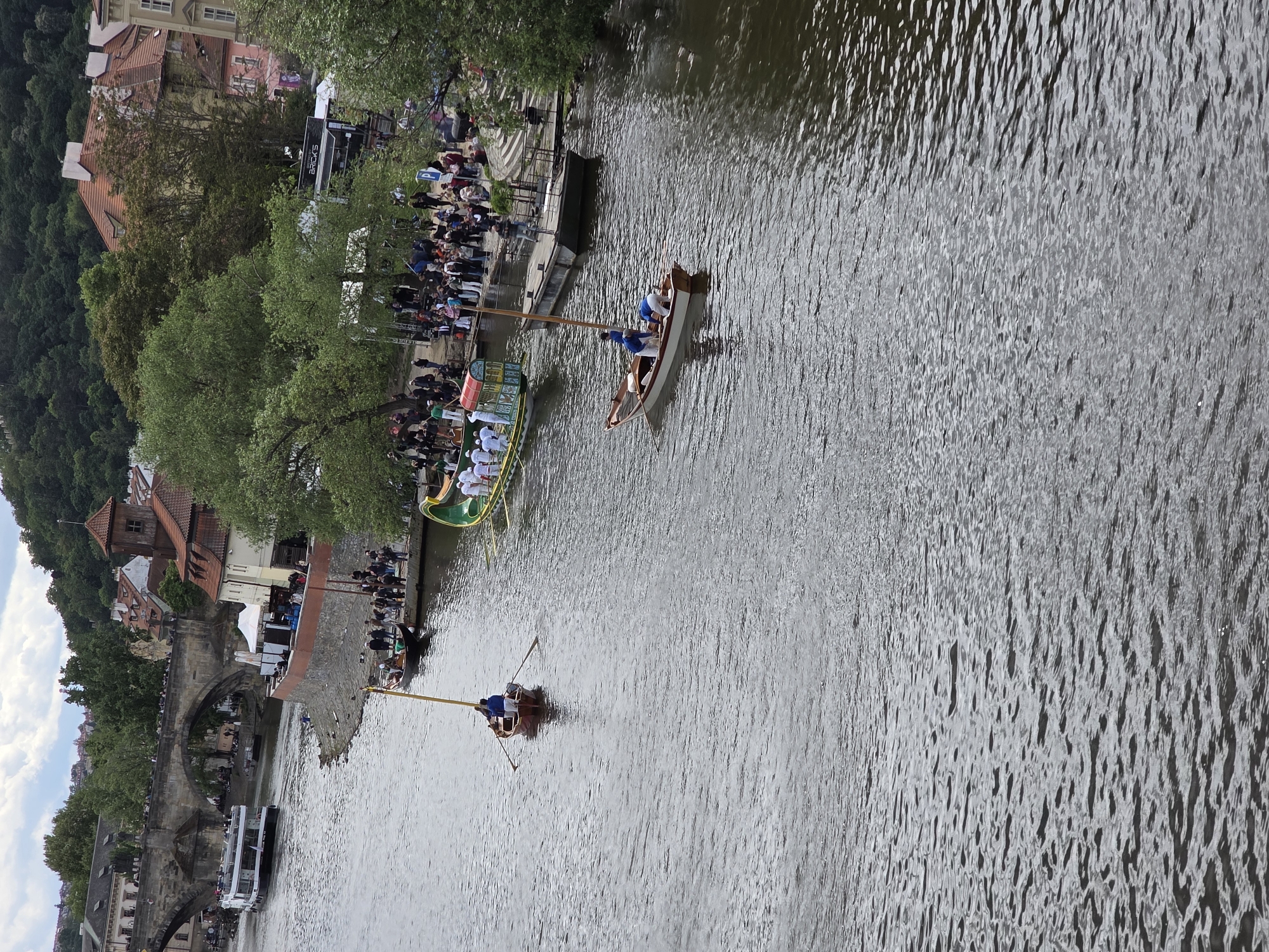 The image captures a river scene in Unknown, Unknown, where two boats are being propelled by individuals using long poles to push off the riverbed. The boats are made of wood with lighter colored interiors, and the people on board are wearing light colored shirts. Crowds of spectators are gathered on the riverbanks, positioned along the edges of a grassy area and a stone walkway. The walkway runs beside the river and has steps leading down to the water level. A long, green and yellow boat is stationed along the riverbank, likely associated with the event.
The river is the primary element in the foreground and stretches into the distance, reflecting the ambient light. A bridge with stone arches spans the river, with buildings adjacent to the bridge, one being a passenger boat, and more people are also visible on the bridge.
The environment includes urban structures intermixed with natural greenery, suggesting a city with historical elements. Trees are present along the riverbank and on the hills beyond the urban development.