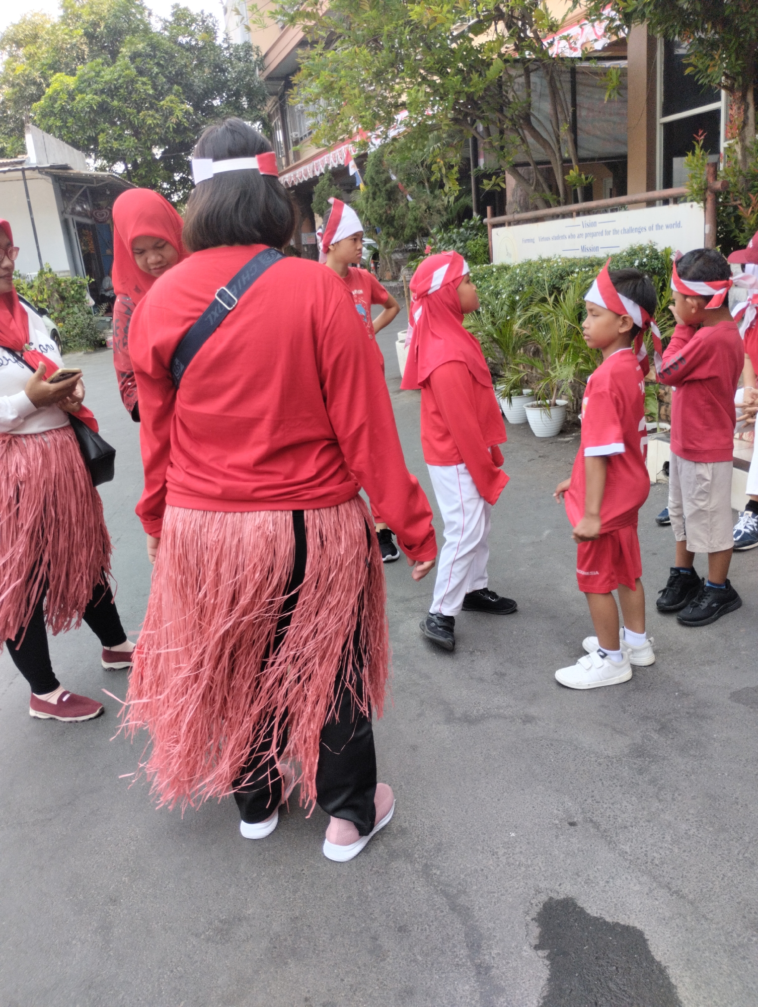 In Bekasi, Indonesia, a group of individuals, predominantly children, are gathered outdoors, likely on a street or paved pathway. The scene is vibrant with the color red, as many participants are wearing red clothing and accessories, including shirts, headbands, and hijabs. Two individuals, possibly adults, are wearing long, tasseled skirts that reach their knees, also in shades of red or pink. The children are dressed in red shirts and shorts or pants, some with red and white headbands, suggesting a patriotic or celebratory event. One child is looking towards the camera, while others are interacting with each other or looking in different directions. The environment features trees, buildings in the background, and planters with greenery. A sign with text is visible on a building, but the content is not fully legible. The overall atmosphere appears to be that of a community gathering or a parade.