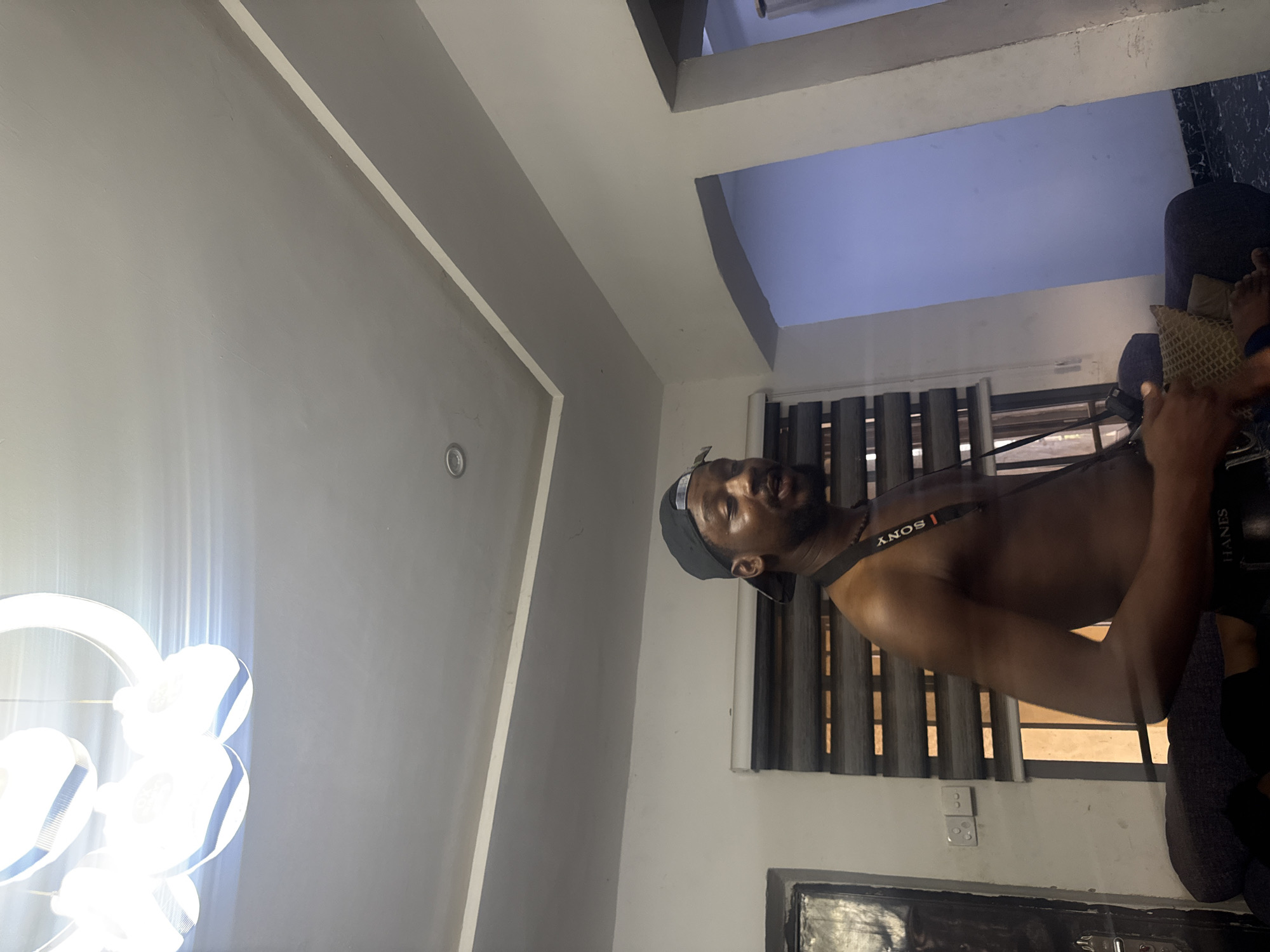 A medium, indoor shot depicts a shirtless, dark-skinned man wearing a black cap and a Sony camera strap. The man is seated in a room in Abuja, Nigeria. Behind him is a window with closed blinds and an electrical outlet on the wall. A modern chandelier is in the top left of the frame. The room has painted grey walls.