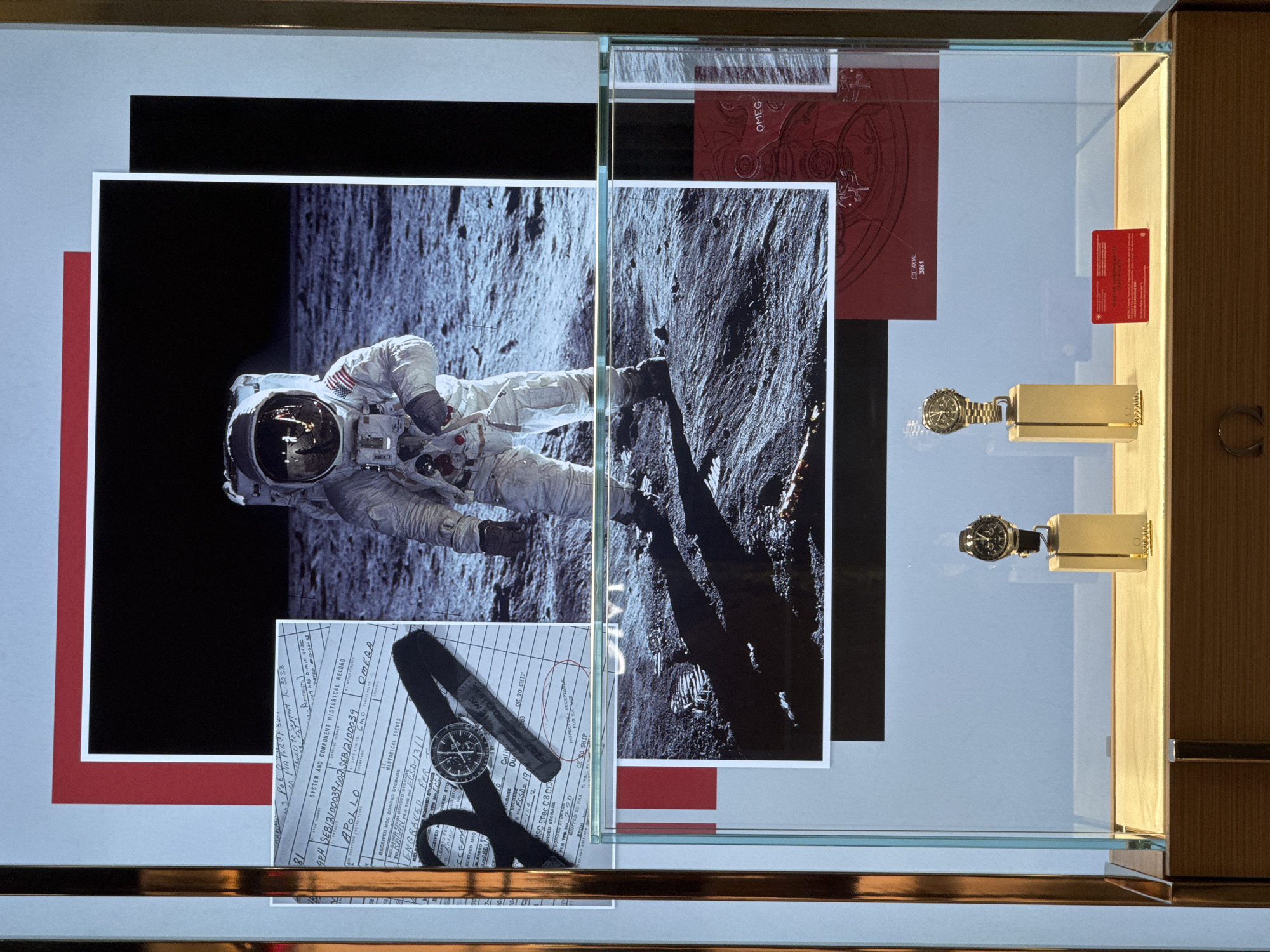 The image shows a window display of a luxury watch store, likely Omega, in Nong Prue Subdistrict, Thailand. The display features a large photograph of an astronaut on the moon, a vintage document with the Apollo mission name, and two Omega watches on stands. The astronaut photograph is prominently placed in the upper half of the display. Below the astronaut image, a document is partially visible, showing handwritten text and details about the Apollo mission and the Omega brand. The lower half of the display showcases two Omega watches, one on a gold-colored stand and another to its right. A red card with text is also visible in the lower right of the display. The entire display is framed by a light-colored border and set within a glass cabinet.