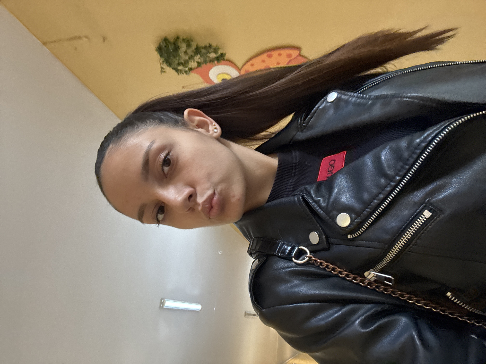 A young woman is photographed in an indoor location. She wears a black leather jacket, a black sweater with a red logo (possibly "Hugo"), and has dark hair pulled back in a ponytail. She is facing the camera with a slight pout. In the background, there is a light yellow ceiling with a cartoonish image of an owl. The other wall is white with a light fixture. The location in Sofia, Bulgaria is unknown, since nothing gives it away.