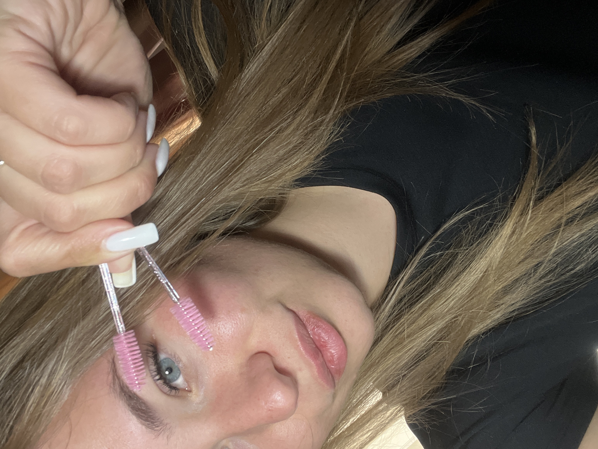 A woman with light brown hair and a black shirt is holding two pink eyelash brushes close to her face. Her fingernails are painted white. The scene appears to be indoors. There is no information suggesting the location is Budva, Montenegro.