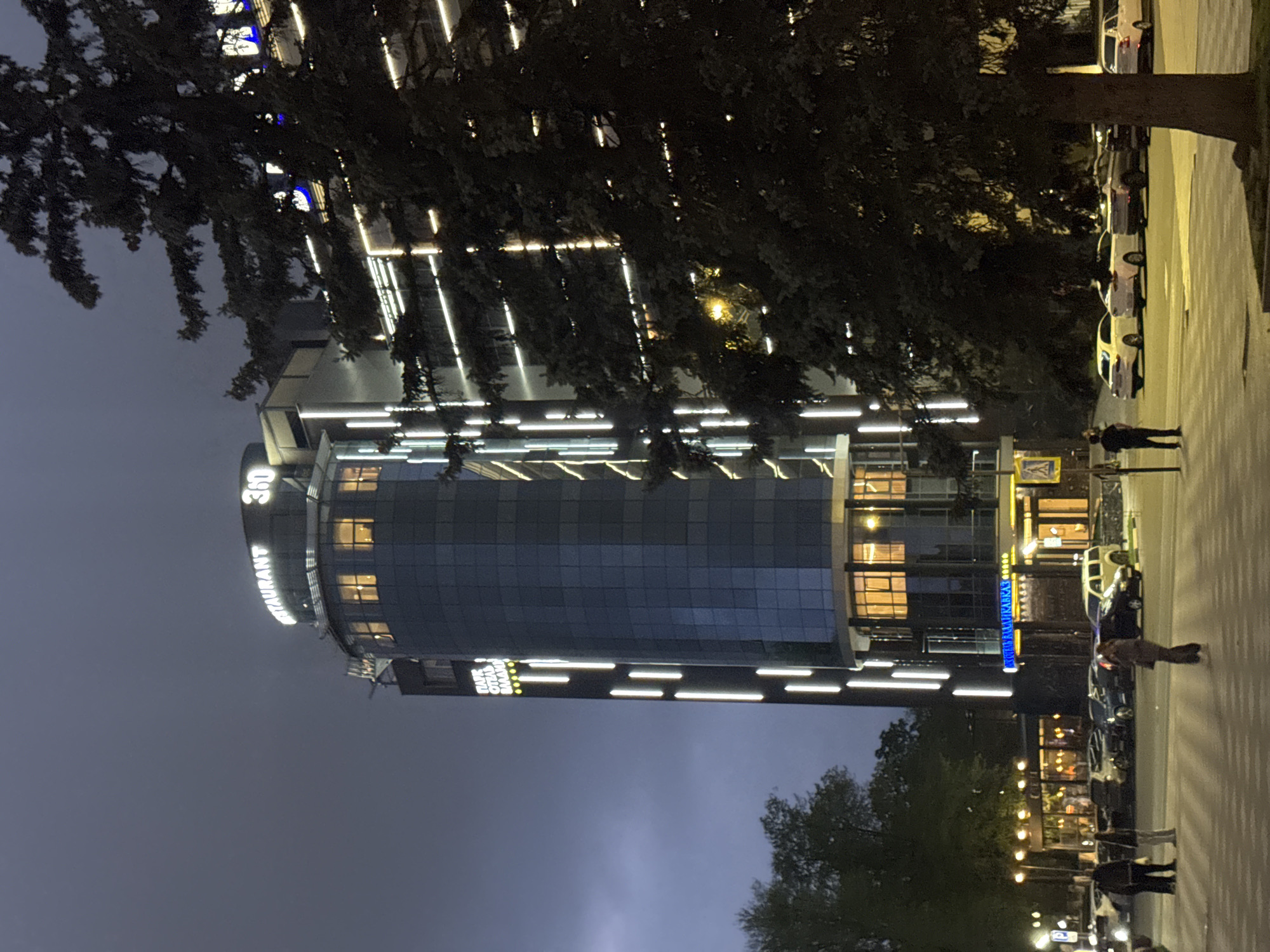The image depicts a tall, modern building with numerous illuminated horizontal lines, likely a hotel or office complex, at night. The building has "360 RESTAURANT" illuminated on its side. Trees are partially obscuring the view of the building. Along the right side of the image is a street with parked cars and pedestrians. The location is Komgaron, Russia.