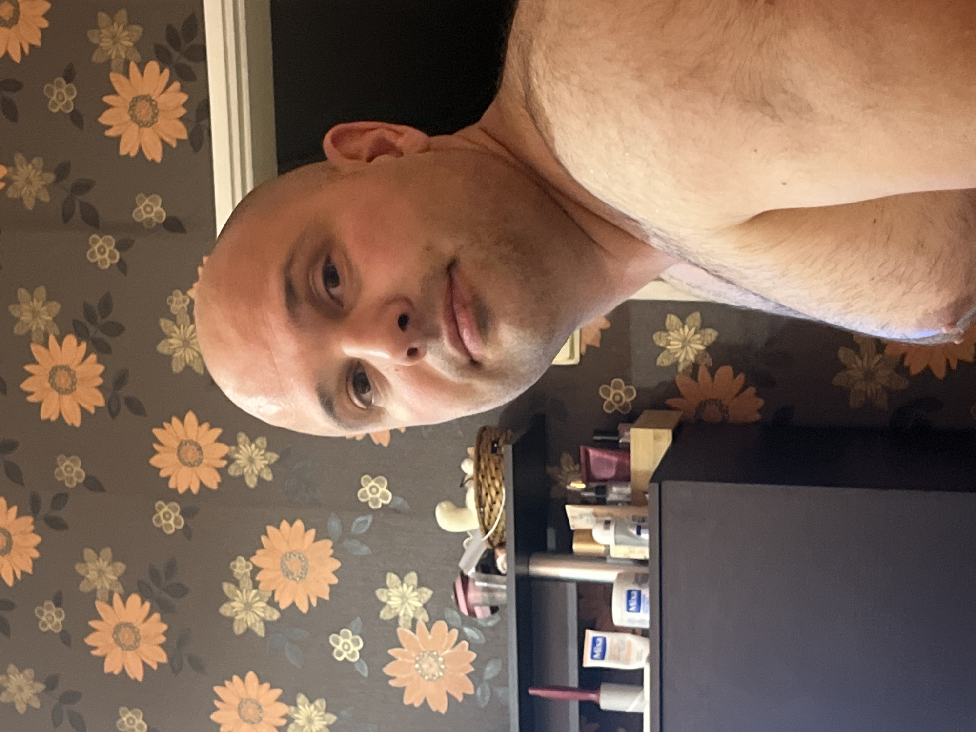 The image is a close-up, slightly overhead shot of a bald, fair-skinned man with a hairy chest and shoulders, looking towards the camera. He has a light stubble on his face and his right ear is visible. The background features a floral wallpaper with orange and yellow flowers on a dark brown base. To the left of the man, a dark grey cabinet is partially visible, with a shelf above it containing several toiletries, including bottles, tubes, and a small woven basket. The scene appears to be indoors, possibly a bathroom or bedroom, but there are no specific indicators of its location within the city of Plovdiv, Bulgaria.