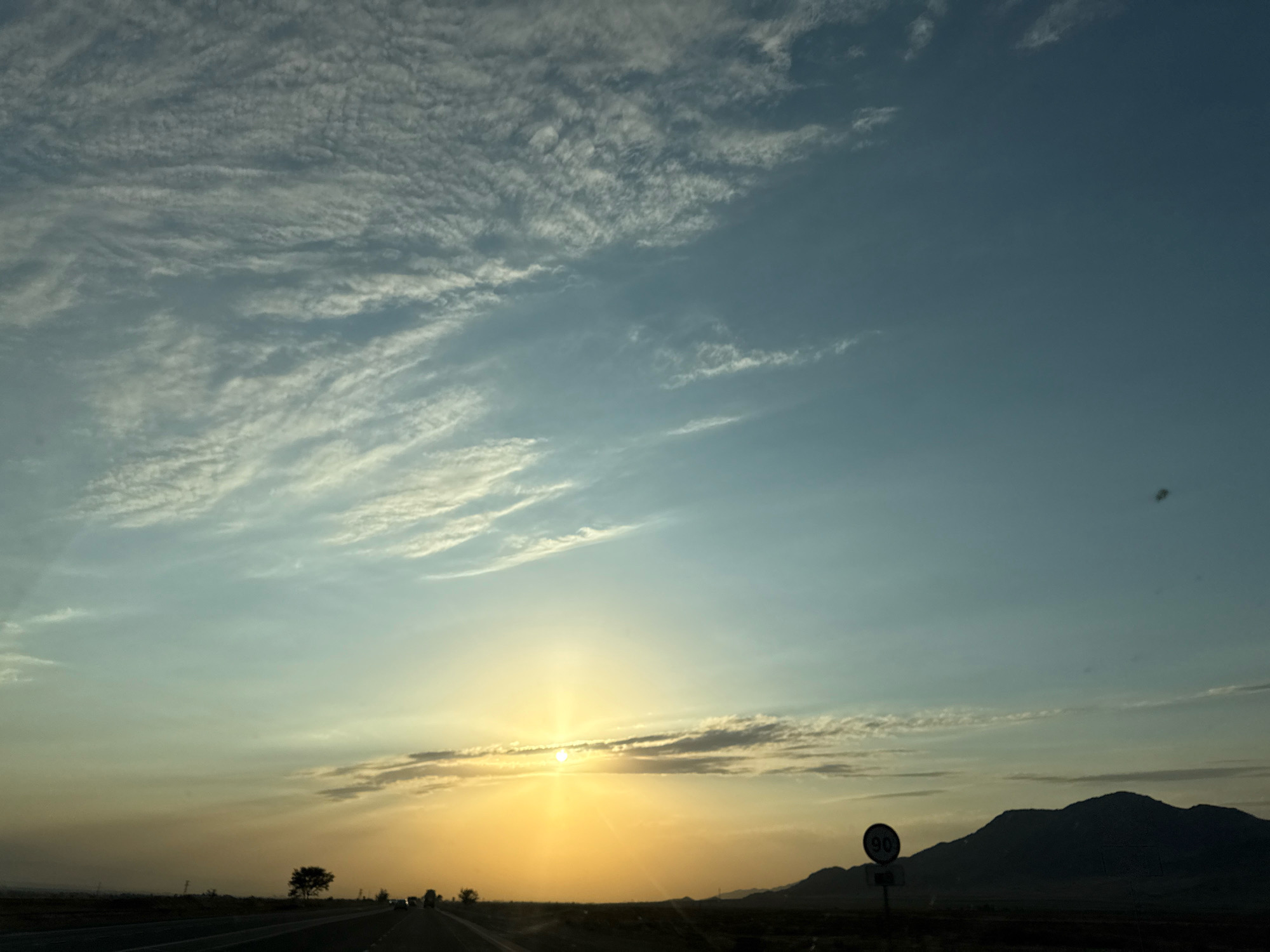 The image depicts a sunset over a highway in the Chüy Region of Kyrgyzstan. The sun is low on the horizon, casting a warm, golden light that illuminates the sky. Wispy clouds are scattered across the sky, tinged with pink and orange from the setting sun. In the foreground, a highway stretches into the distance, with a few cars visible. To the right, a speed limit sign indicating "90" is posted. In the far distance, a silhouette of mountains can be seen against the colorful sky. A lone tree stands on the side of the road. The overall scene is one of natural beauty and tranquility.