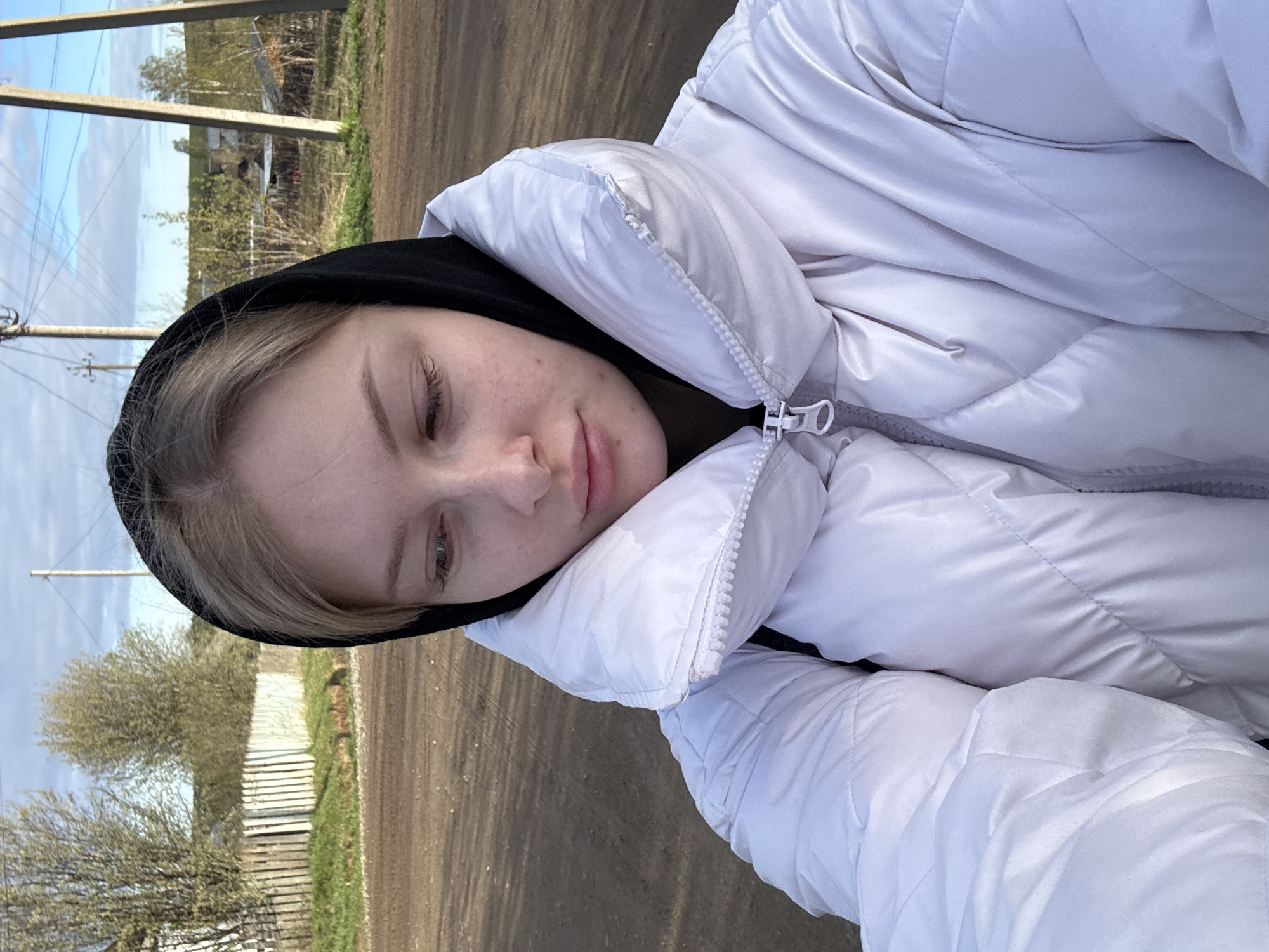 A medium shot photograph captures a young, fair-skinned woman wearing a light-colored puffer jacket with a black hood. She is lying down, with her head slightly tilted. Her eyes are partially closed. The background reveals an outdoor scene, with a dirt road visible. The presence of telephone poles and a fence suggests a rural or suburban area. The setting is in Бородулино, Russia.