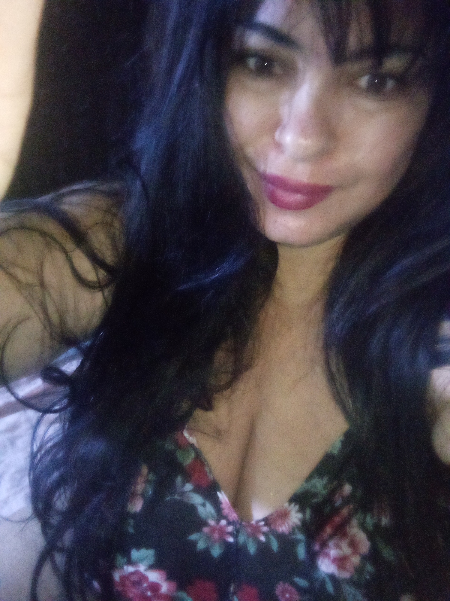 A medium shot of a woman with long, dark hair and bangs, wearing a floral-patterned dress. Her lips are painted with a dark pink lipstick. The background is dark and indistinct. The location is unknown.