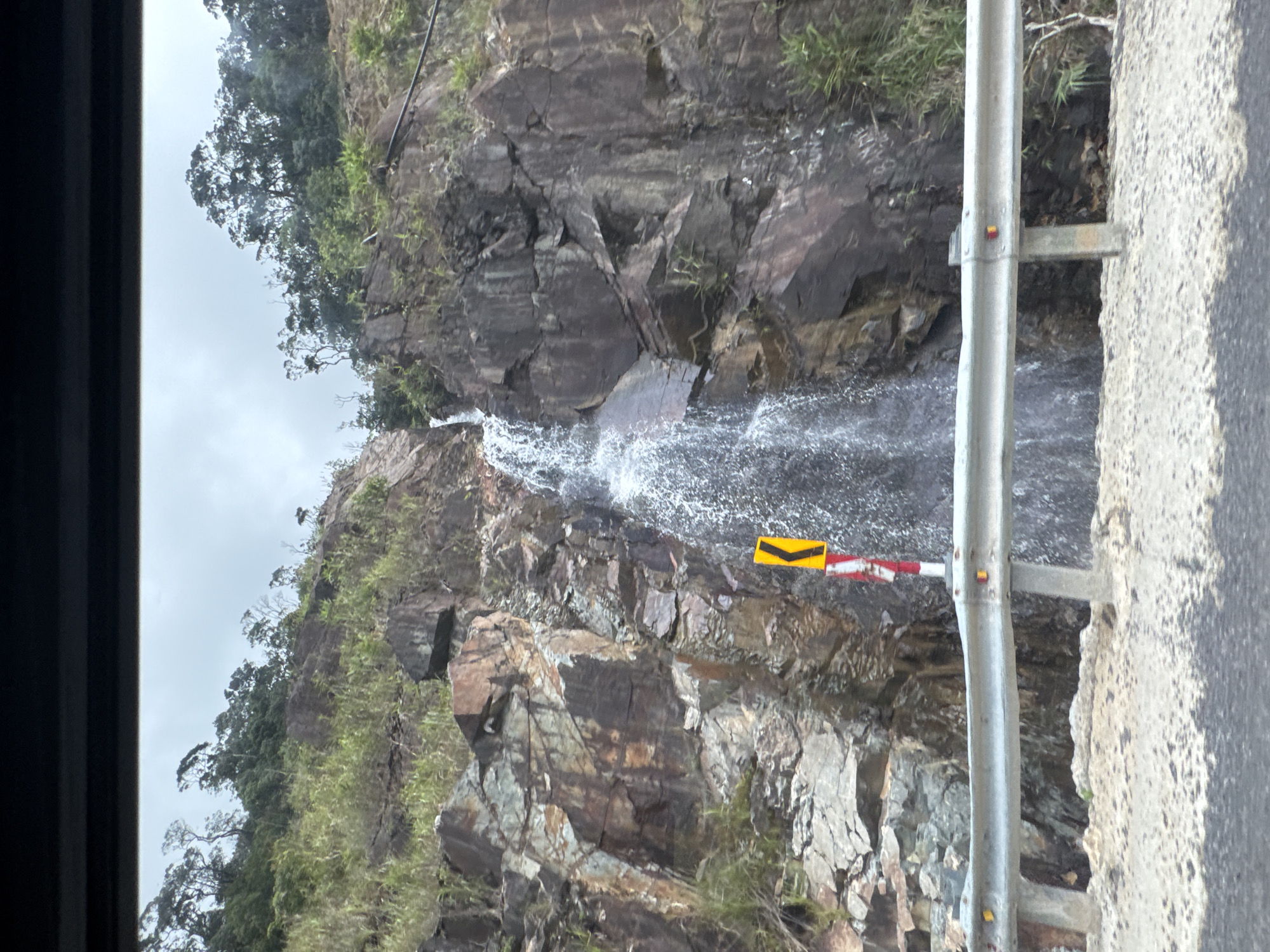 The image captures a scenic view of a waterfall cascading down a rocky mountainside, possibly along a roadside in Nam Khanh Vinh, Vietnam. The waterfall is the central focus, with water flowing over jagged brown and grey rocks. Green vegetation, including grasses and trees, grows on the slopes of the mountain, contrasting with the rocky terrain.
In the foreground, a metal guardrail runs horizontally across the frame, indicating the presence of a road. The guardrail is silver and appears sturdy, with some yellow and red reflective markers. Beyond the guardrail, a portion of the road surface is visible, appearing to be paved with asphalt and gravel.
A yellow chevron sign with a black arrow pointing left is mounted on a pole beside the waterfall, suggesting a curve in the road or a hazard. The sky above is overcast and a pale grey, hinting at cloudy weather. The overall scene depicts a natural landscape with elements of infrastructure, suggesting it's a location accessible by road. There are no people or animals visible in the image.