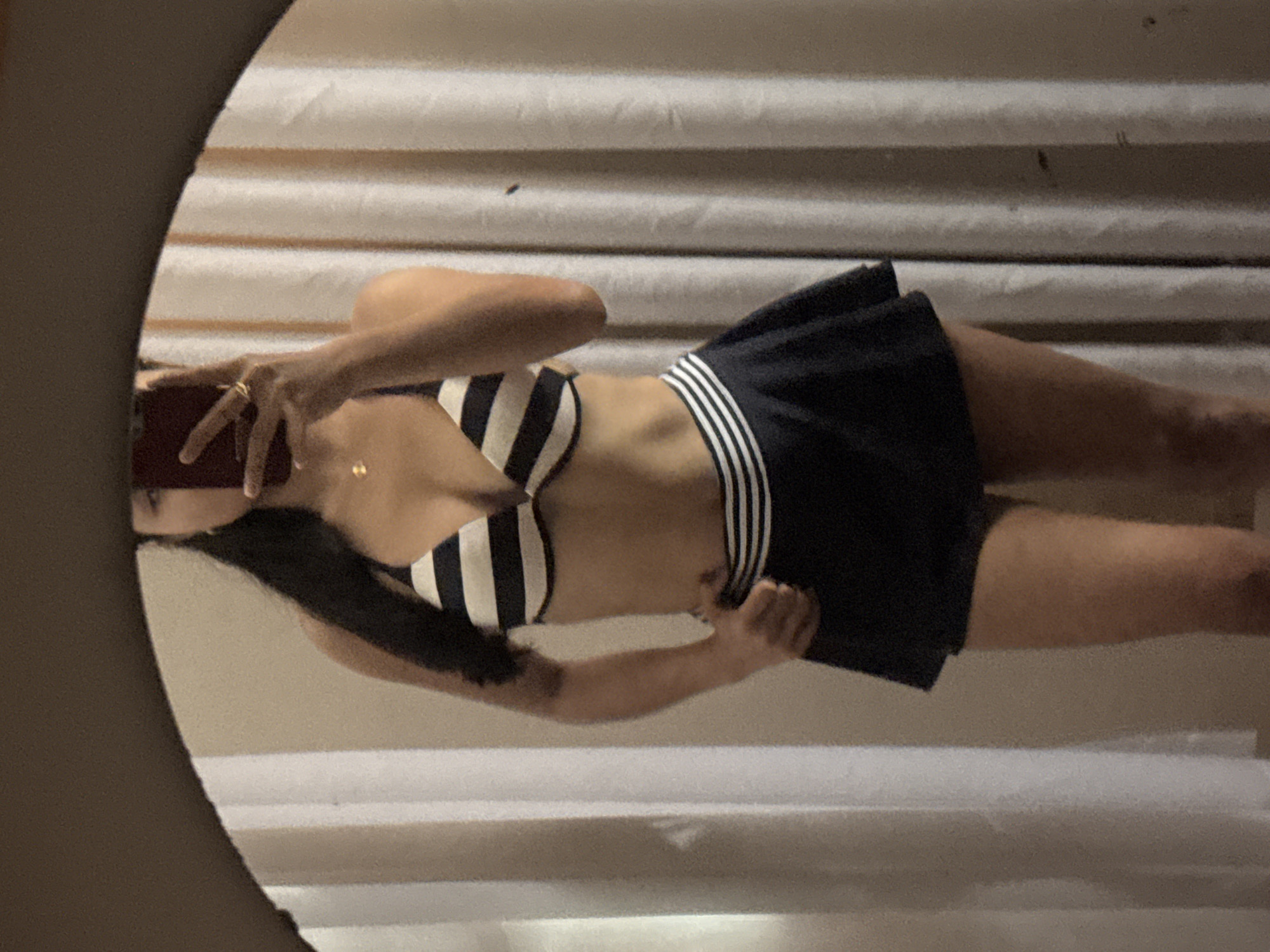 An individual, partially visible from the chest down and with their face obscured by a dark-colored smartphone, is captured in a mirror selfie taken within what appears to be a private room in Davao City, Philippines. The person possesses long, dark hair draped over their left shoulder and is wearing a two-piece swimwear set. The bikini top features horizontal black and white stripes, while the bottom is a dark blue or black swim skirt with a distinct white and dark striped waistband. A golden ring is present on the person's right ring finger, and a delicate gold pendant necklace hangs from their neck. The individual's left hand rests on their left hip. The background, visible in the mirror's reflection, consists of light-colored curtains with vertical folds, covering a wall of a neutral tone. The mirror itself has a dark, solid frame visible along the top edge of the image. The lighting is consistent with an indoor setting.