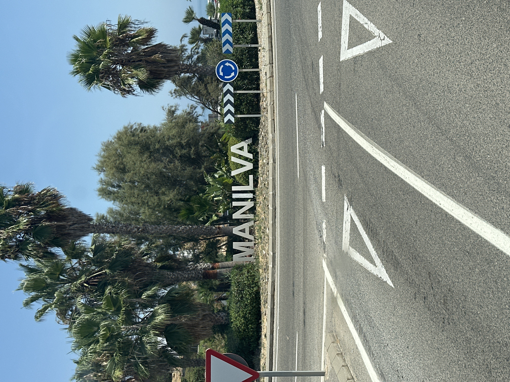 The image depicts a street scene in Manilva, La Chullera, Spain. In the foreground, a road surface is visible with lane markings, including yield triangles painted in white. Behind the road, a large sign with white, three-dimensional letters spells out "MANILVA." Surrounding the sign is landscaping with green bushes and gravel. Several palm trees are prominent in the background, along with other trees and foliage. Road signs are present, including directional chevron signs and a circular blue sign indicating a roundabout. The sky is clear and blue. The overall impression is of a sunny day at a roadside entrance to the town.