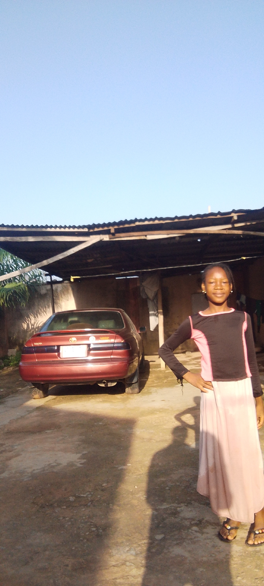 A young girl stands in front of a red car parked under a corrugated metal awning. The girl is wearing a black long-sleeved shirt with pink accents on the shoulders and sides, and a long, light pink skirt. She has her right hand on her hip and is smiling at the camera. The car is a sedan with a spoiler on the trunk and a license plate that is not clearly visible. The awning is made of corrugated metal sheets supported by wooden beams and appears to be part of an open-air structure, possibly a carport or an outdoor workshop. In the background, there is a building with a rough texture on its walls. The ground is a dirt or gravel surface. The sky is clear and blue, suggesting it is daytime. The sunlight casts long shadows on the ground, indicating it might be late afternoon or early morning. The overall scene appears to be in a rural or semi-urban setting. There are no other people clearly visible in the image, though a shadow of a person taking the photo is cast on the ground. The girl's expression is happy and relaxed.