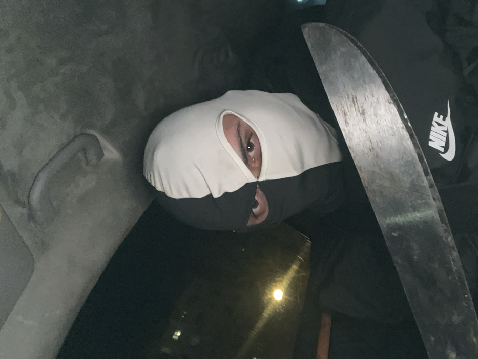 An individual is photographed in a close-up from within a vehicle, situated in the city of Plovdiv, Bulgaria, during nighttime. The person is wearing a two-toned balaclava, vertically split with the left half (viewer's left) black and the right half white, revealing only their dark brown eyes. They are dressed in a dark garment, possibly a sweatshirt, displaying a white "NIKE" logo on the right chest area. A large, broad-bladed metallic knife, resembling a machete, is held horizontally across the lower part of the frame, partially obscuring the individual's torso. The blade exhibits minor wear and surface imperfections. The vehicle's light-colored headliner and an overhead grab handle are visible in the upper frame. A distinct, circular light source from outside the vehicle, along with vague building silhouettes, is visible through the window on the left, indicating the nocturnal setting.