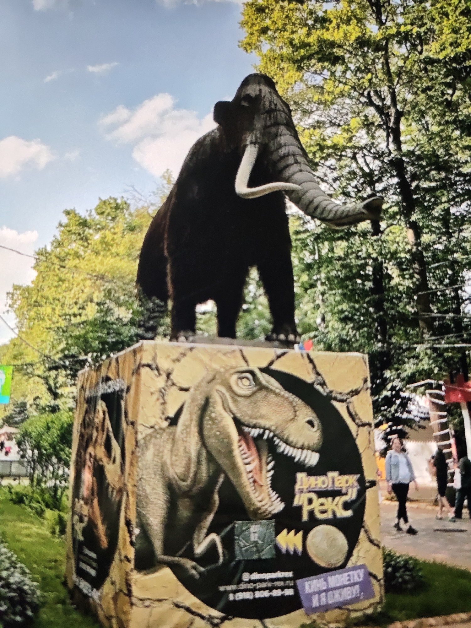 A statue of a mammoth stands on a pedestal decorated with images of dinosaurs. The pedestal has text in Russian that reads "ДиноПарк 'Рекс'" and "КИнь МОНЕТКУ И Я оживУ!" The text indicates the location is Dinopark "Reks" in Stavropol, Russia. In the background, trees and a blue sky with clouds are visible. Several people are walking in the background.