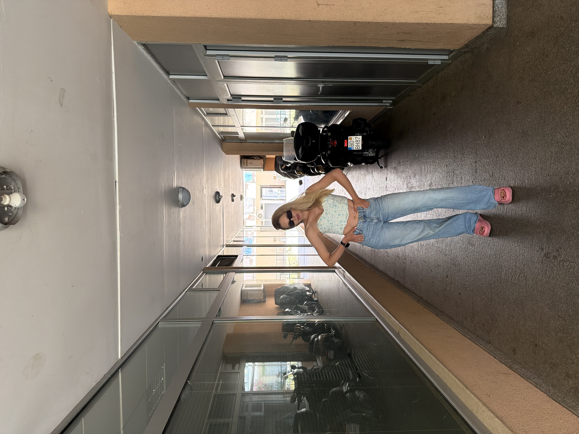 A blonde woman stands centered in an outdoor covered passageway, hands on her hips, looking directly at the viewer. She wears black sunglasses, a light blue and white floral strapless crop top, light blue wide-leg jeans, and pink open-toed slides. A dark-colored watch is visible on her left wrist. The passageway features a textured concrete floor, light peach/orange stucco-textured walls, and a light grey ceiling with multiple surface-mounted light fixtures and visible stains.
On the left side of the passageway, a continuous glass wall, framed in dark grey metal, reflects a series of parked motorcycles or scooters indoors, along with interior building details and exterior views. On the right side, a solid peach-colored wall transitions to a section with a dark grey framed glass door and additional glass panels. A black scooter with a large black top case is parked along this wall; its rear license plate "BD BH87" is clearly visible, identifying Budva, Montenegro as the location. Beyond the parked scooter, reflections in the glass further reveal exterior buildings and foliage. Natural light illuminates the scene, casting shadows on the floor and walls.