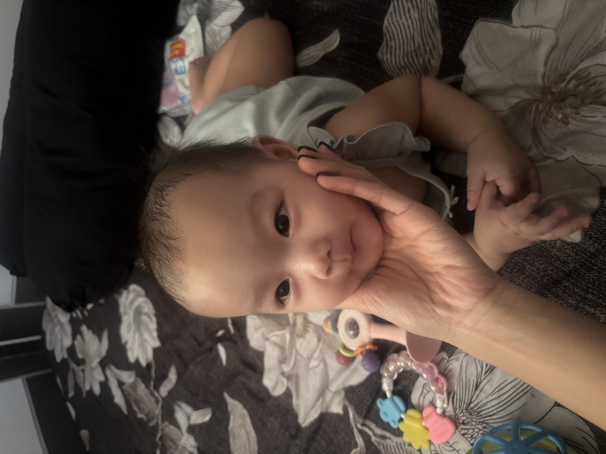 A medium shot captures a baby lying on a patterned sheet. The baby, facing left, is being supported by a hand cupping its cheek. The baby's head is tilted slightly, and its eyes are open and looking towards the viewer. Toys, including a rattle and a colorful teething ring, are scattered around the baby. The background consists of a dark, patterned sheet and a blurred black pillow. The scene appears to be indoors, possibly in a bedroom, and the overall impression is one of domesticity and care. There is no identifiable information about the specific location within West Jakarta, Indonesia, or any other details that can be inferred from the image alone.