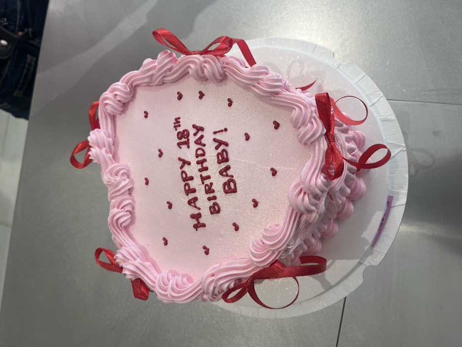 An overhead view captures a heart-shaped cake resting on a white corrugated cardboard cake board, which is placed on a reflective metallic-gray surface. The cake is frosted in light pink with a subtle shimmer and features a piped border of ruffled pink icing around its perimeter. Several small red satin ribbons, tied into bows, are attached to this border. The top surface of the cake is inscribed with "HAPPY 18th BIRTHDAY BABY!" in red capital letters and is further embellished with small, scattered red heart-shaped sprinkles. A small purple label is visible beneath the cake board on the left side. In the upper right background, a partial view of a person's lap or upper thigh is visible, showing dark blue fabric, a dark belt, and a silver-colored buckle. The specific location within Paris, France, cannot be determined from the visual evidence.