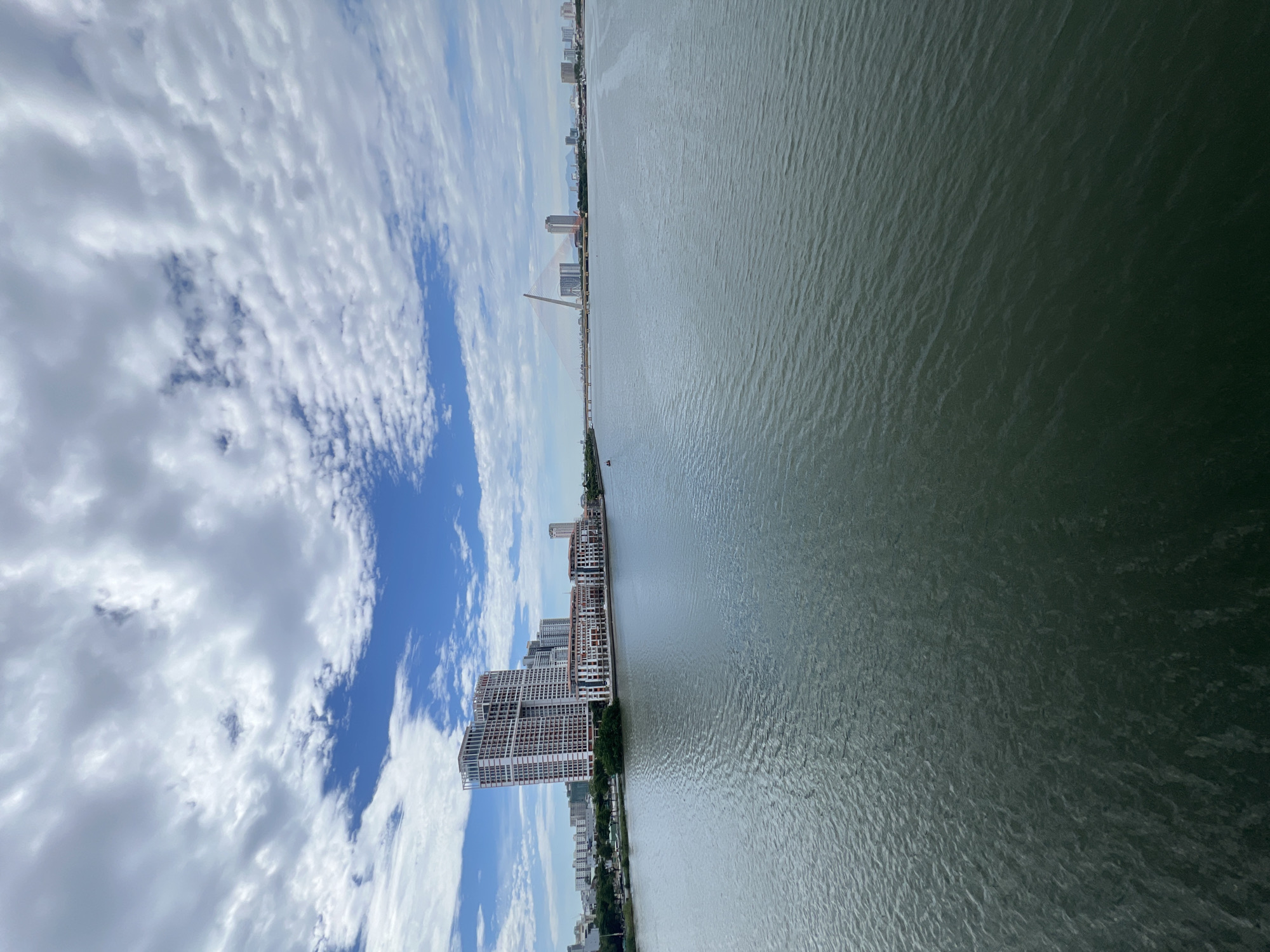 The image displays a broad view of the Han River and its surrounding urban landscape in Đà Nẵng, Vietnam, captured on a brightly lit day. The sky, occupying the upper portion of the frame, features a mix of clear blue and numerous scattered white cumulus clouds. On the left bank of the river, a prominent, multi-story high-rise building with a white and reddish-brown exterior and complex architectural recesses dominates the immediate cityscape. Adjacent to and in front of this tower are lower-rise buildings exhibiting a similar design, lining the river's edge. Behind these structures, a dense array of smaller urban buildings and some green foliage is visible. The wide river, occupying the lower half of the image, presents a dark, greenish-blue surface covered in small, consistent ripples, indicating water movement. A small, dark, indistinct object, possibly a boat, is faintly visible on the river in the mid-ground, towards the left. In the far distance, on the right side of the river, a distinctive cable-stayed bridge with prominent orange-red cables spans the waterway. Beyond the bridge, a distant city skyline composed of various high-rise buildings and urban structures extends along the far bank, with faint outlines of mountains or hills visible on the horizon. No individuals are discernible within the scene.