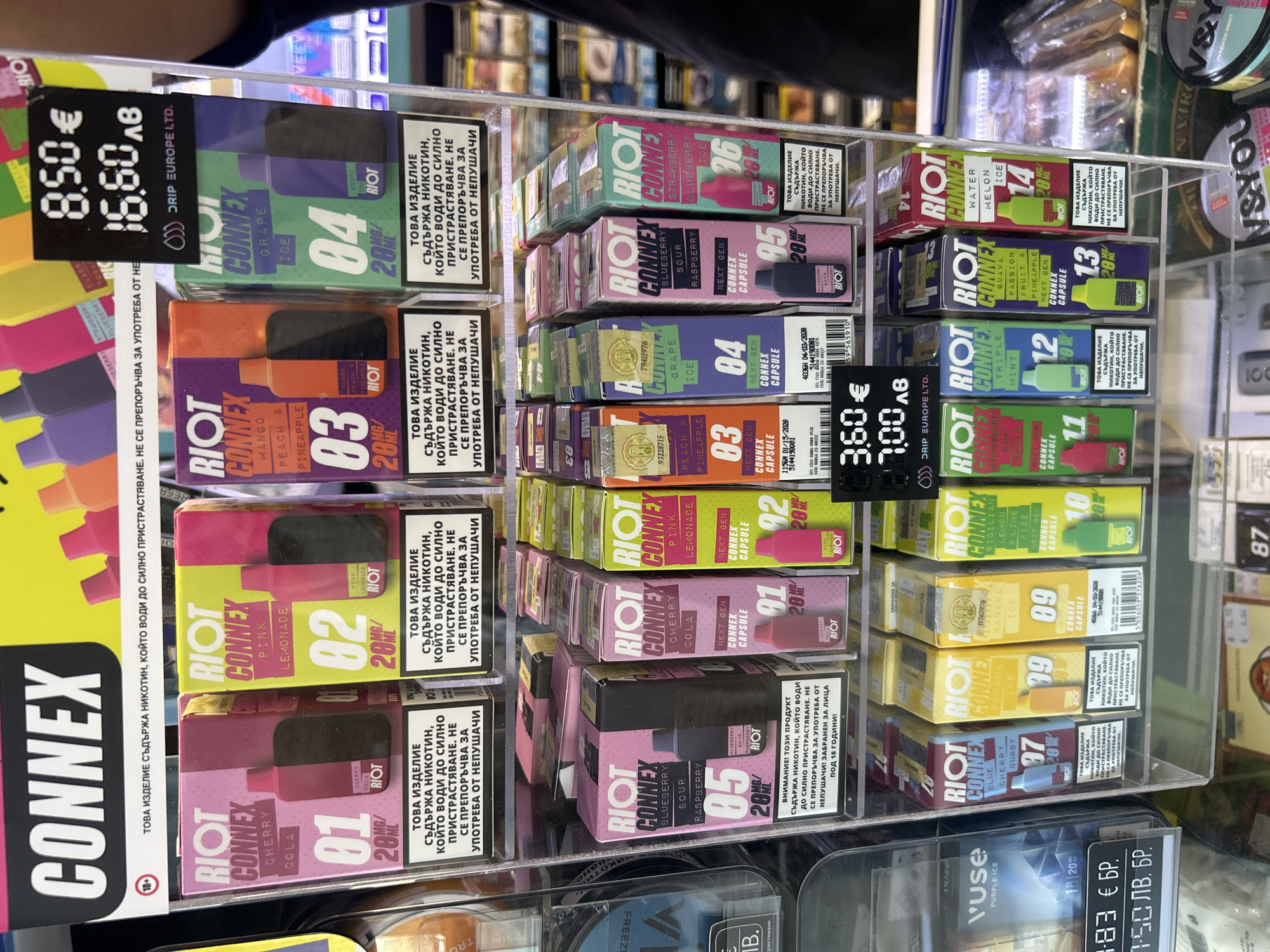 A close-up shot shows a display of vape products in a retail setting. The products are arranged in clear plastic compartments, with each item packaged in a colorful box. The brand "RIOT CONNEX" is prominently featured on many of the boxes, which are color-coded and numbered from 01 to 18, indicating different flavors such as Cherry Cola, Pink Lemonade, Mango Peach Pineapple, Grape Ice, Blueberry Sour Raspberry, and Triple Mint. Some boxes also display the nicotine strength, "20MG/".
Visible text includes the brand name, flavor names, product numbers, nicotine strength, and warning labels in Bulgarian that translate to "THIS PRODUCT CONTAINS NICOTINE, WHICH IS HIGHLY ADDICTIVE. NOT RECOMMENDED FOR USE BY NON-SMOKERS. FORBIDDEN FOR PERSONS UNDER 18 YEARS OF AGE!". There are also price tags displaying prices in Euros and Bulgarian Leva, such as "8.50€ 16.80лв" and "3.60€ 7.00 лв", and "1.83 € БР. 7.50 лв. БР.". Some boxes have manufacturing dates and codes printed on them.
The background is blurred, but appears to be a retail store with shelves of other products, possibly cigarettes or other vaping supplies. The lighting suggests an indoor environment, likely under artificial store lights. There are no people visible in the frame. The overall impression is one of a commercial display designed to showcase a variety of vape products to potential customers.