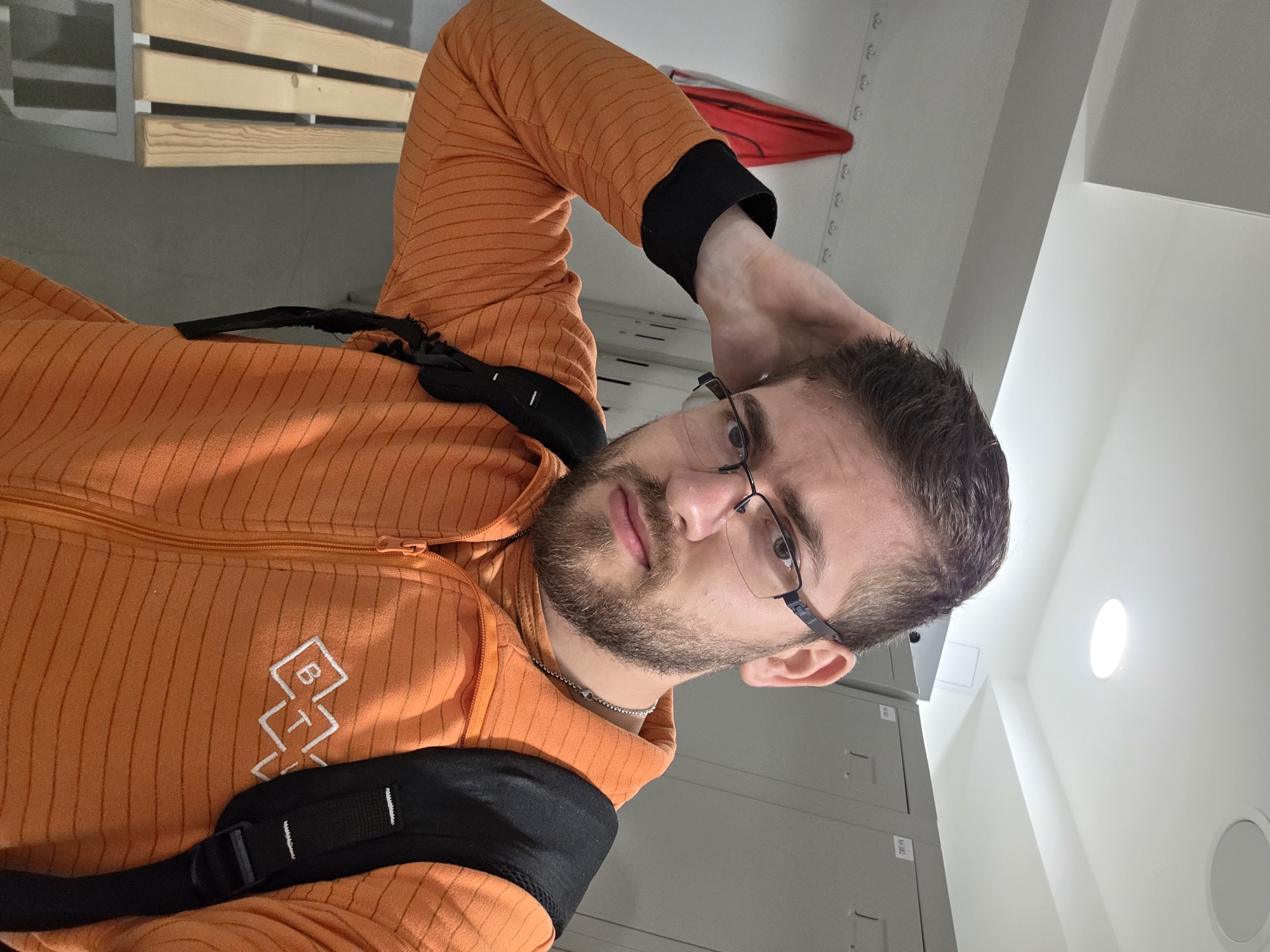 A man with a beard and glasses is indoors, wearing an orange, striped jacket with a black backpack strap across his shoulder. He is looking at the camera with his hand resting on his head. In the background are grey lockers and a white ceiling with a circular light fixture. Part of a wooden bench can be seen in the upper left corner. There is no information to specify the image was taken in Plovdiv, Bulgaria.