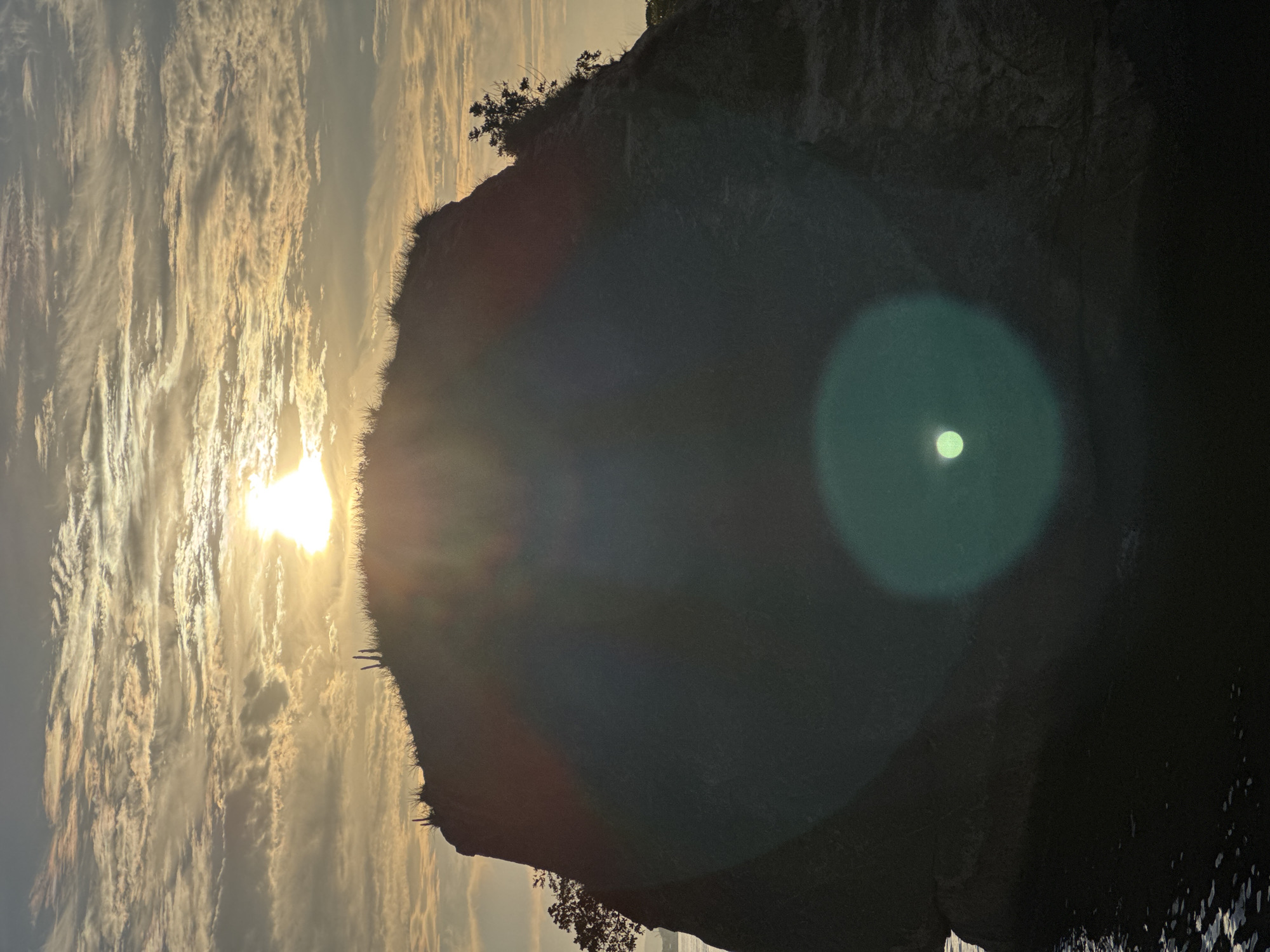 A silhouetted rocky outcrop with sparse vegetation on its summit is partially illuminated by the setting or rising sun. The sun is positioned behind the outcrop, creating a lens flare effect in the foreground. The sky is filled with scattered clouds, with the sun peeking through them. The lower portion of the image shows dark water, likely part of Guanabara Bay. The location is Itacoatiara beach, Niterói, Brazil.