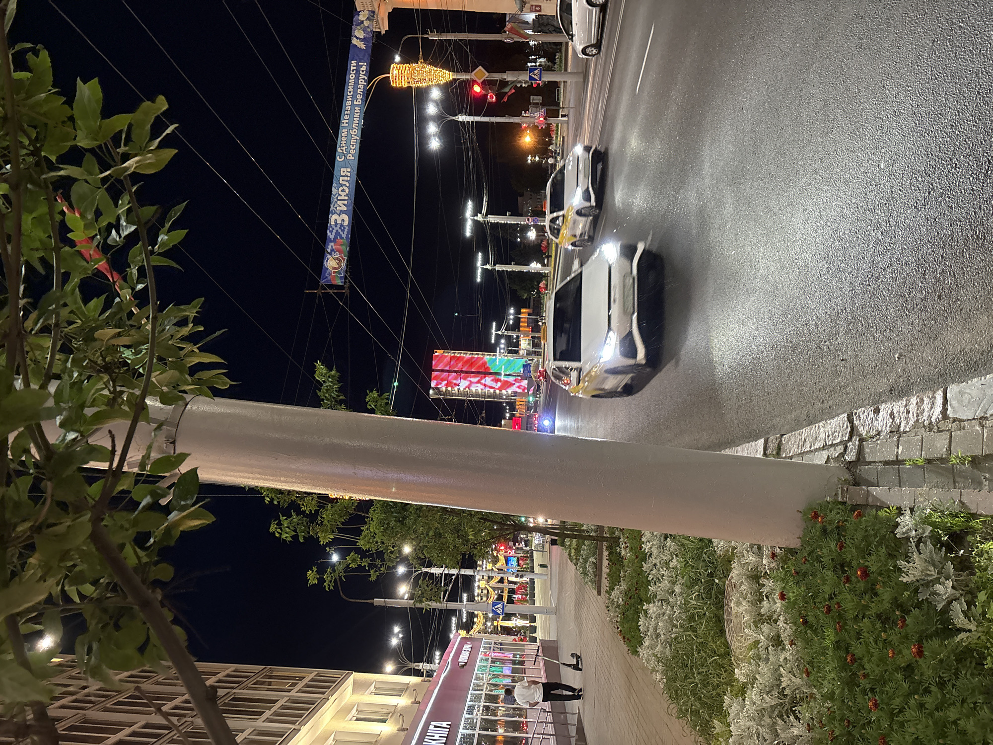 The image depicts a nighttime street scene in Vitsebsk, Belarus, likely on July 3rd, given the banner visible in the upper portion of the frame that reads "С Днём Независимости Республики Беларусь!" which translates to "Happy Independence Day of the Republic of Belarus!".
In the foreground, a white, cylindrical utility pole occupies a significant portion of the right side of the frame. To the left of the pole, a sidewalk runs alongside a street. A man wearing a white t-shirt and black pants is walking on the sidewalk, pulling a black electric scooter behind him. Adjacent to the sidewalk, a planter bed contains green foliage and small red flowers. Further down the sidewalk, a building with a red facade and large windows is visible, with illuminated lettering on its front.
The street itself is wet, reflecting the lights from the surrounding environment. Several cars are present on the road. A white sedan with its headlights illuminated is driving towards the viewer in the foreground. Behind it, a white car with yellow accents, possibly a taxi, is also in motion. Further in the distance, traffic lights and street lamps illuminate the scene. A large digital screen displaying red and green colors is visible on the right side of the street in the background. Overhead, a banner hangs across the street with the aforementioned Independence Day greeting. The sky is dark, indicating it is nighttime.