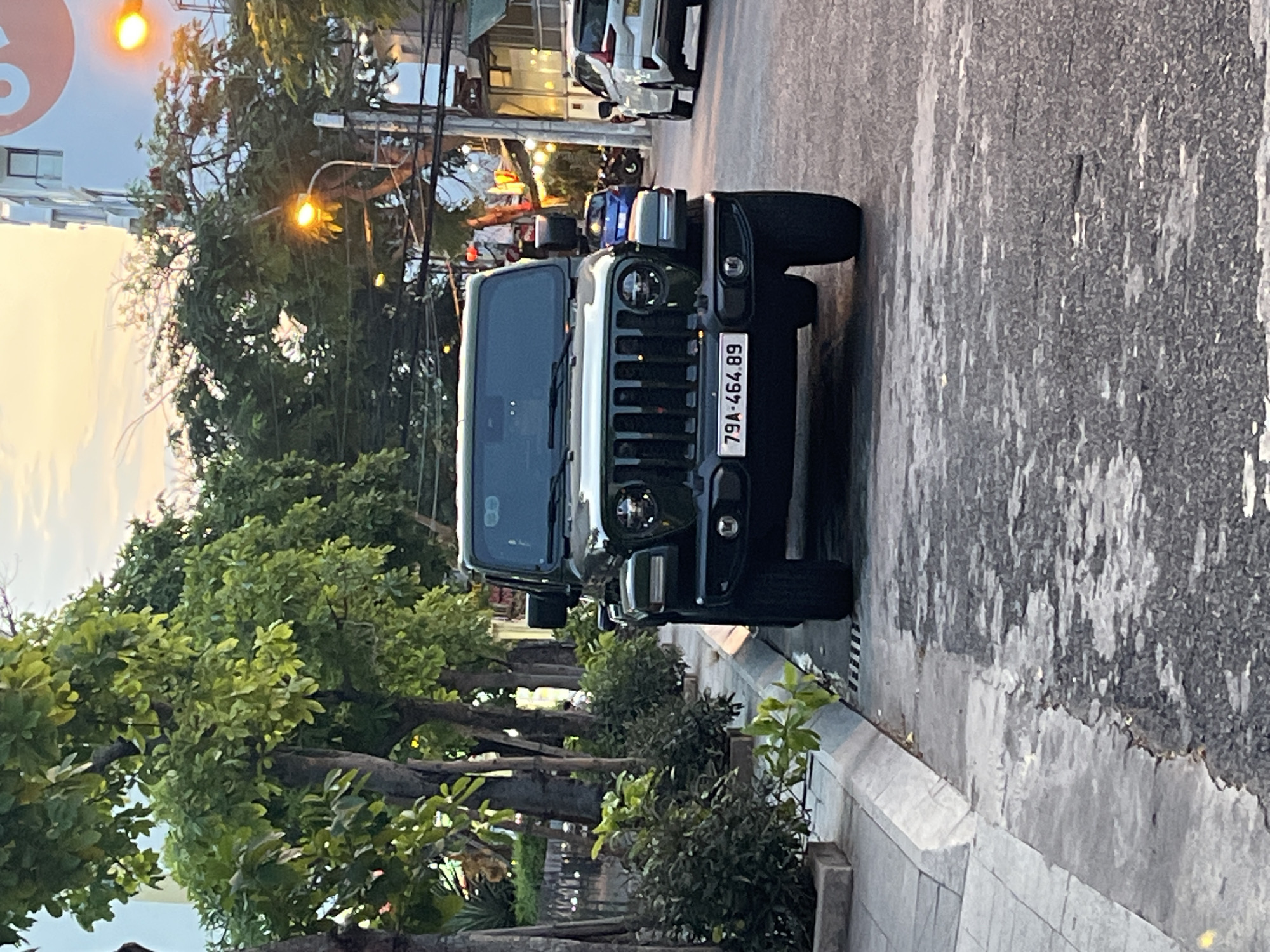 The image shows a black Jeep parked on the side of a street, with the license plate visible, reading "79A-464.89." The street is lined with trees and bushes. In the background, there are buildings and other vehicles, including a white car and a possible police vehicle with blue lights. The scene appears to be in an urban area, specifically Bac Nha Trang Ward, Vietnam. The road surface is asphalt, showing signs of wear and patching. The sky is overcast, with a soft light filtering through.