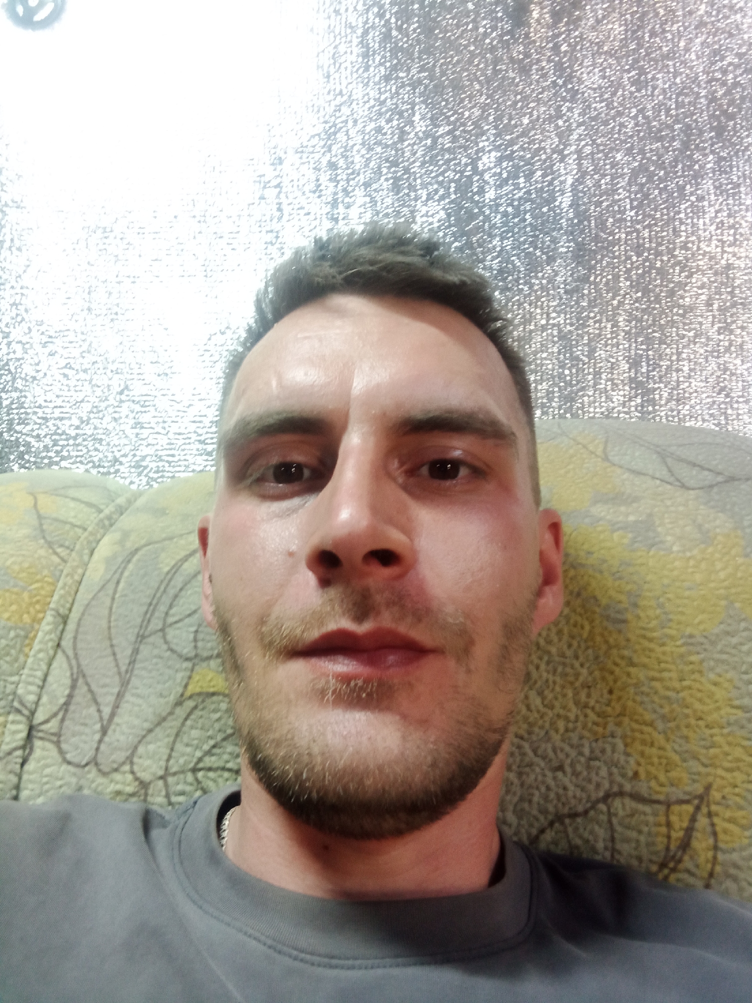 A close-up selfie shows a man with short, dark hair and a stubble beard. He is wearing a grey sweatshirt and a silver chain is visible around his neck. The background consists of a textured surface that could be a wall or ceiling, with an irregular speckled pattern. Behind him is a patterned couch with beige and yellow tones, and brown branches. The location of the photo is Chaikovsky, Russia.