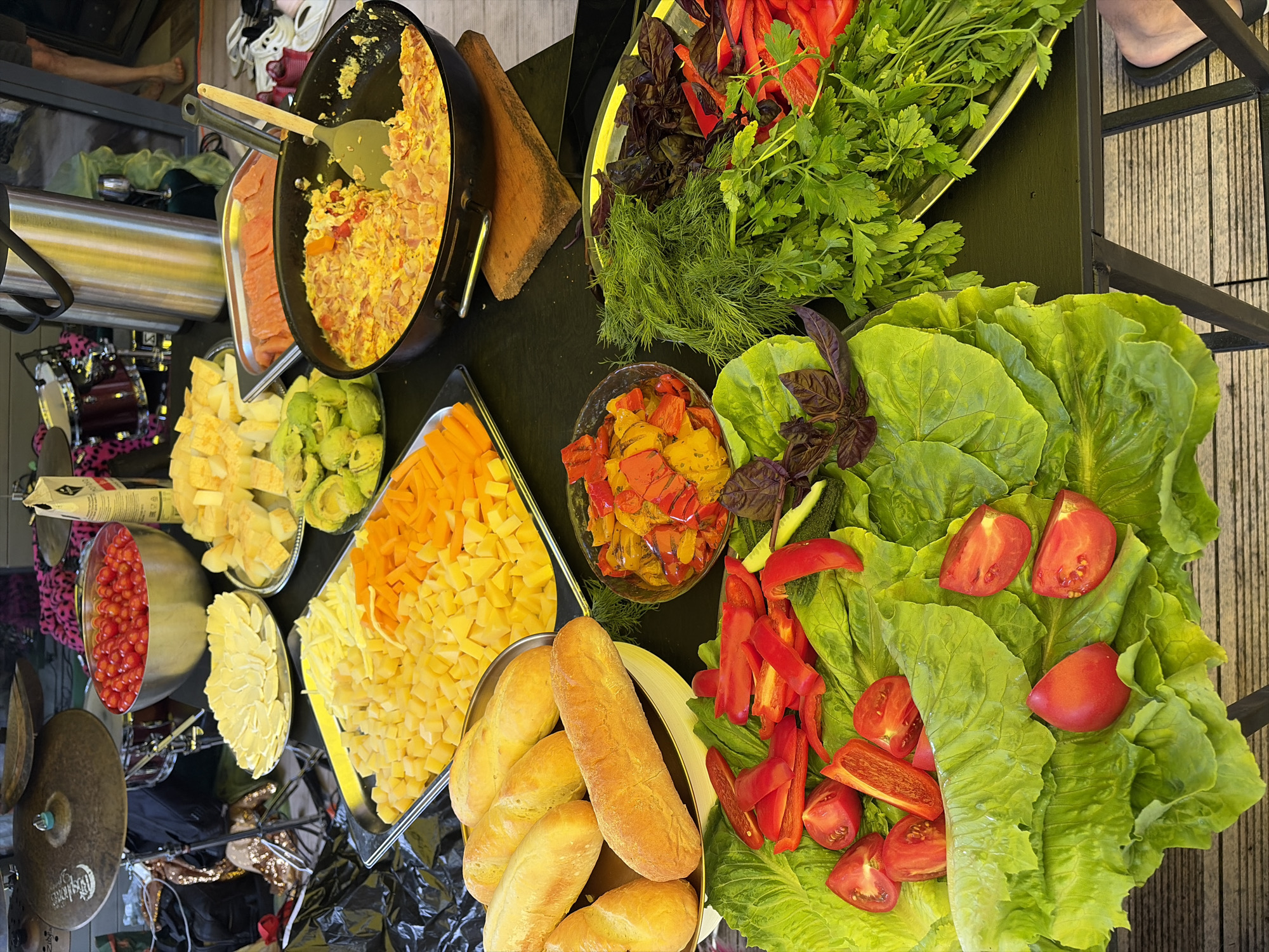 An outdoor food spread is laid out on a dark table, likely in Shebanovo, Russia. In the foreground, a large platter of vibrant green lettuce leaves is topped with halved cherry tomatoes and sliced red bell peppers. Adjacent to this, a tray holds a generous assortment of fresh herbs, including dill and parsley, along with purple basil leaves and more sliced red bell peppers. To the right, a bowl contains cooked vegetables such as peppers and onions. Further back, a pan holds a cooked egg dish, possibly scrambled eggs with bacon, resting on a wooden board. A separate tray displays thin slices of salmon. Other items on the table include several loaves of bread, cubed cheeses in yellow and orange hues, and a bowl filled with small red tomatoes. In the background, parts of a drum set are visible, along with a large stainless steel thermos or beverage dispenser and various bags and fabrics with animal prints. The scene is illuminated by natural light.