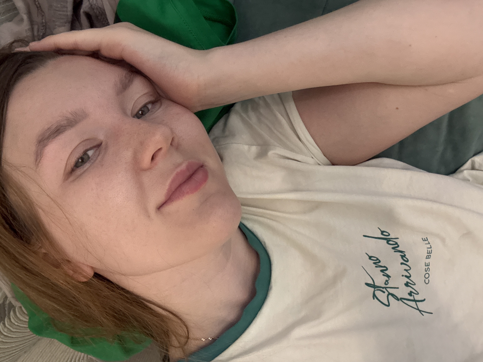 A young, light-skinned woman with light brown hair is lying down. She is wearing a light-colored t-shirt with green trim and green text that reads "Stanno Arrivando COSE BELLE". Her left arm is raised, with her hand resting near her forehead. The background is soft and indistinct, with hints of green and other muted colors. There is no information to suggest a location in the city of Izhevsk, Russia.