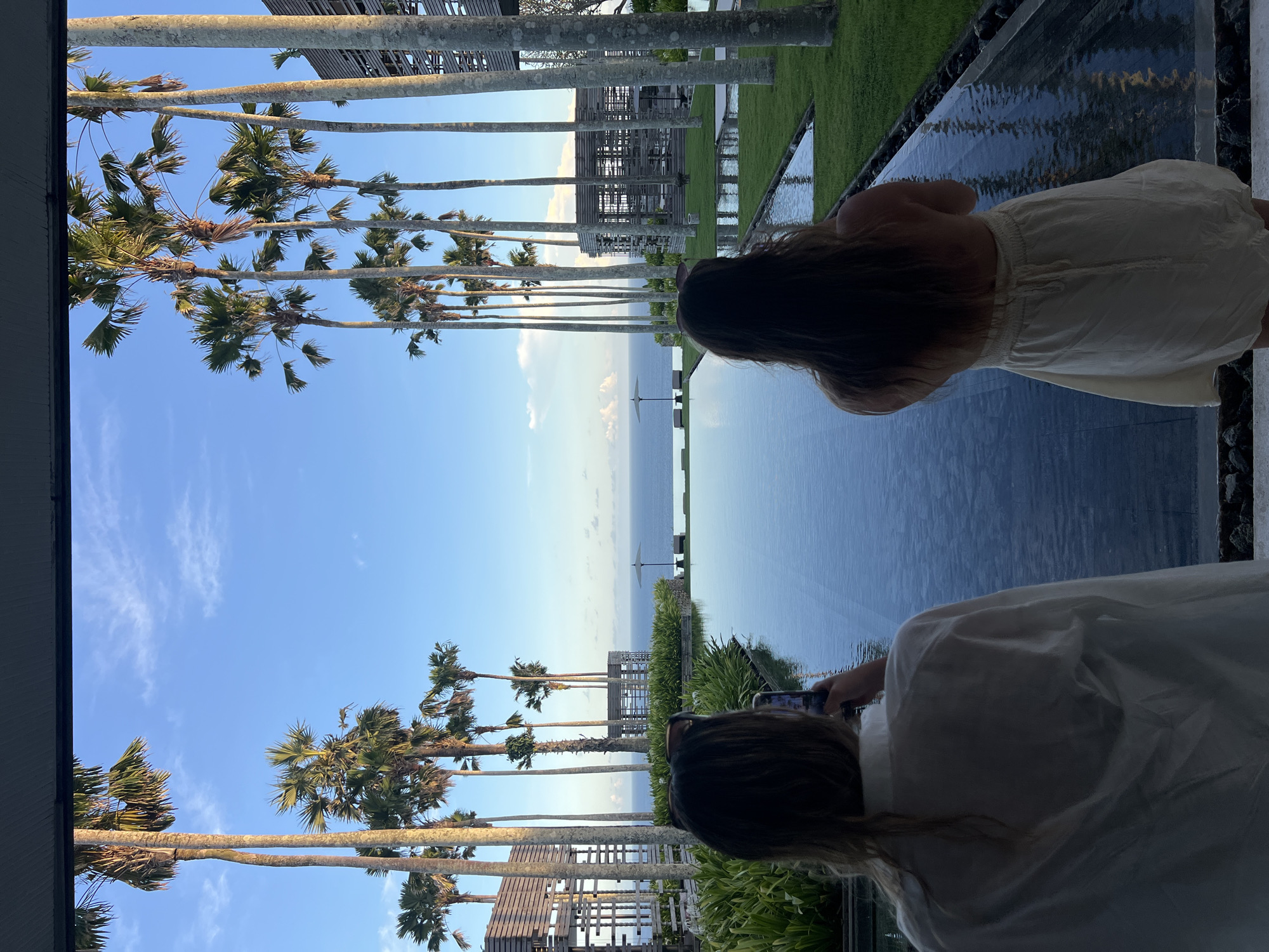 This image presents a serene tropical resort scene, looking out towards the ocean from a raised perspective in Kuta Selatan, Indonesia. The setting appears to be late afternoon or early evening, characterized by warm light and a clear sky.
In the immediate foreground, with their backs to the camera, are two individuals. On the left, a person wearing a white shirt is holding a smartphone, seemingly engaged in photographing or videoing the expansive view. To their right, slightly further back, another person dressed in a light-colored skirt or dress stands, also observing the scenic landscape.
They are positioned beside a wide, dark-tiled infinity pool or decorative water feature that reflects the sky and distant elements. Beyond this water feature, a prominent row of tall palm trees with slender trunks extends horizontally across the mid-ground, creating a natural frame for the ocean beyond. Amidst and behind the palm trees, several open-air, modern resort structures are visible. The background features the vast, calm expanse of the ocean meeting a clear blue sky, which is dotted with scattered, delicate white clouds. A solitary white sun umbrella can be discerned far out on the water, near the horizon.
The overall impression conveyed is one of peaceful observation and appreciation of a beautiful coastal landscape, typical of a luxurious tropical getaway. There is no visible text in the image.
