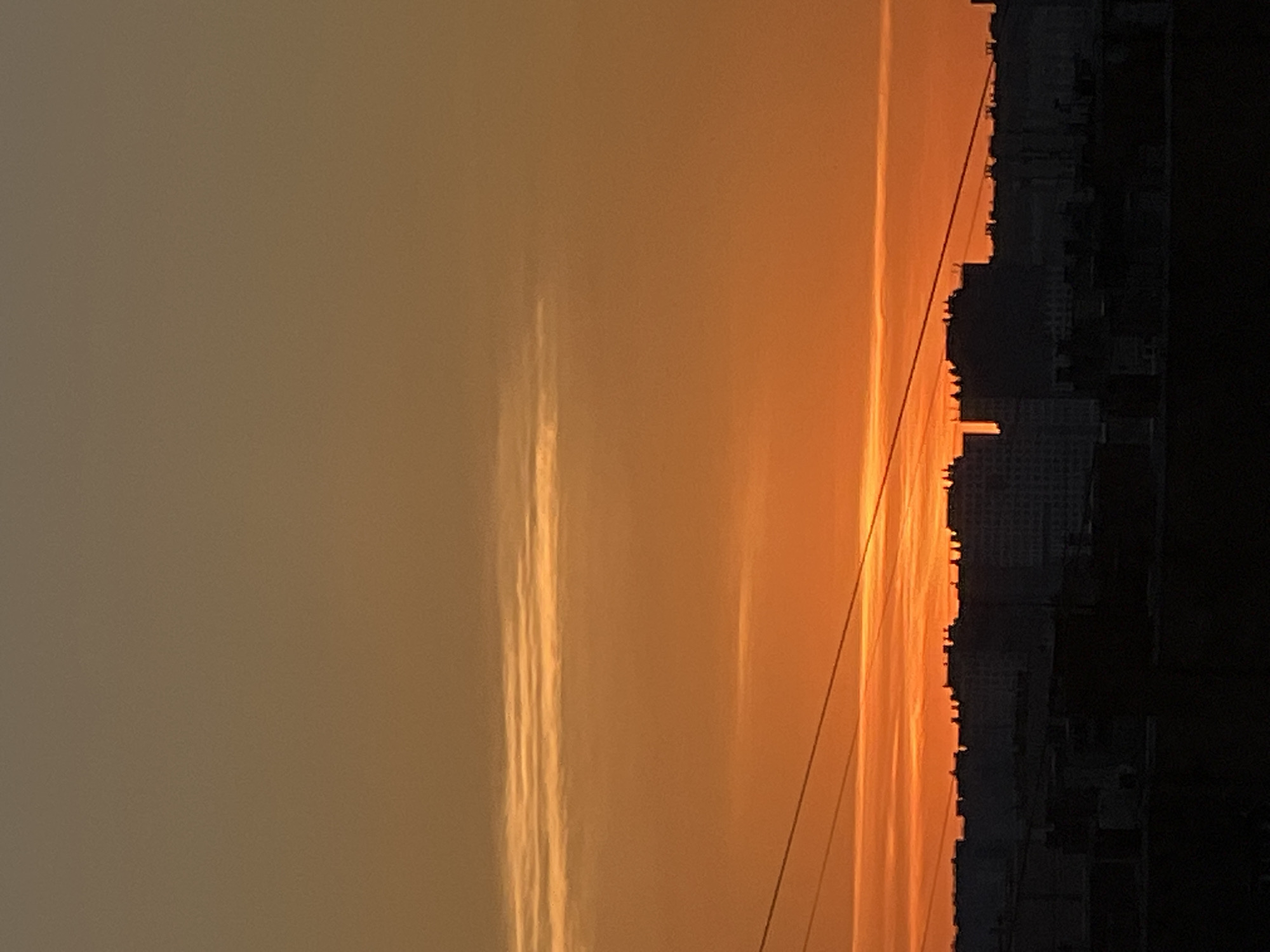 The image depicts a sunset over the city of Sinyavino, Russia. The sky is a gradient of orange and yellow, with thin, horizontal clouds streaking across the upper half. Silhouetted against the vibrant sky is the dark outline of a cityscape, featuring the rooftops of numerous buildings. Several thin, dark lines, likely power lines, run diagonally across the frame, adding a graphic element to the scene. The lower portion of the image is dominated by the dark silhouettes of foreground buildings and structures, obscuring finer details.