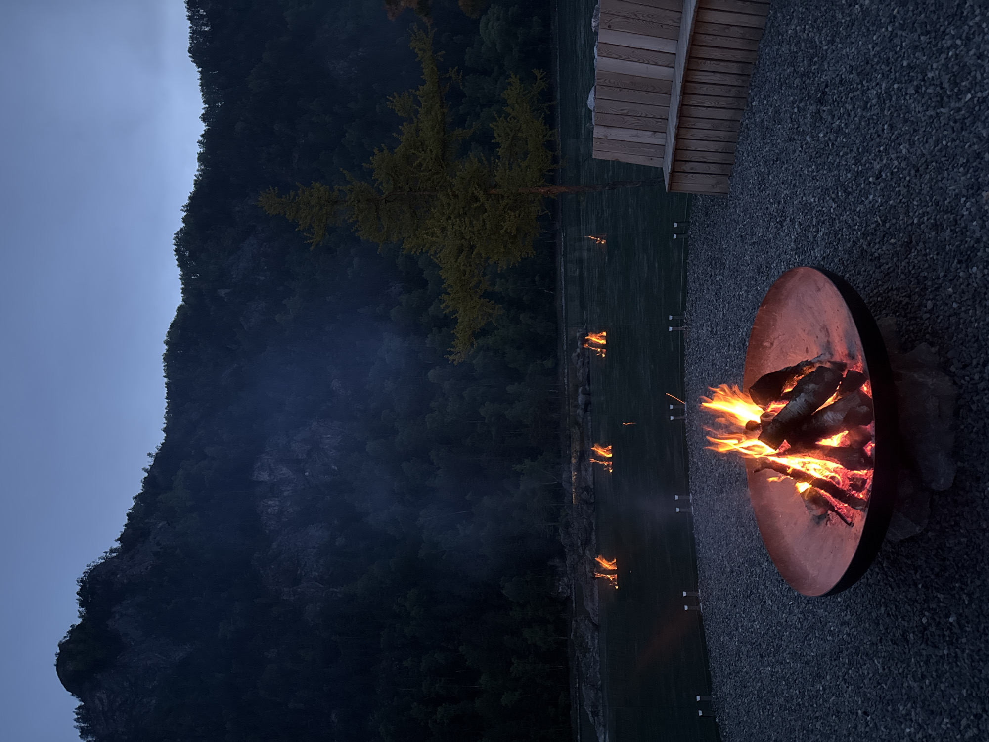 A fire pit with burning wood stands on a gravel surface in Ust'-Muny, Russia. In the background is a steep, tree-covered mountain, and a line of flames appears to be located on the lake. A wooden structure is partially visible in the upper right corner. The sky is overcast.