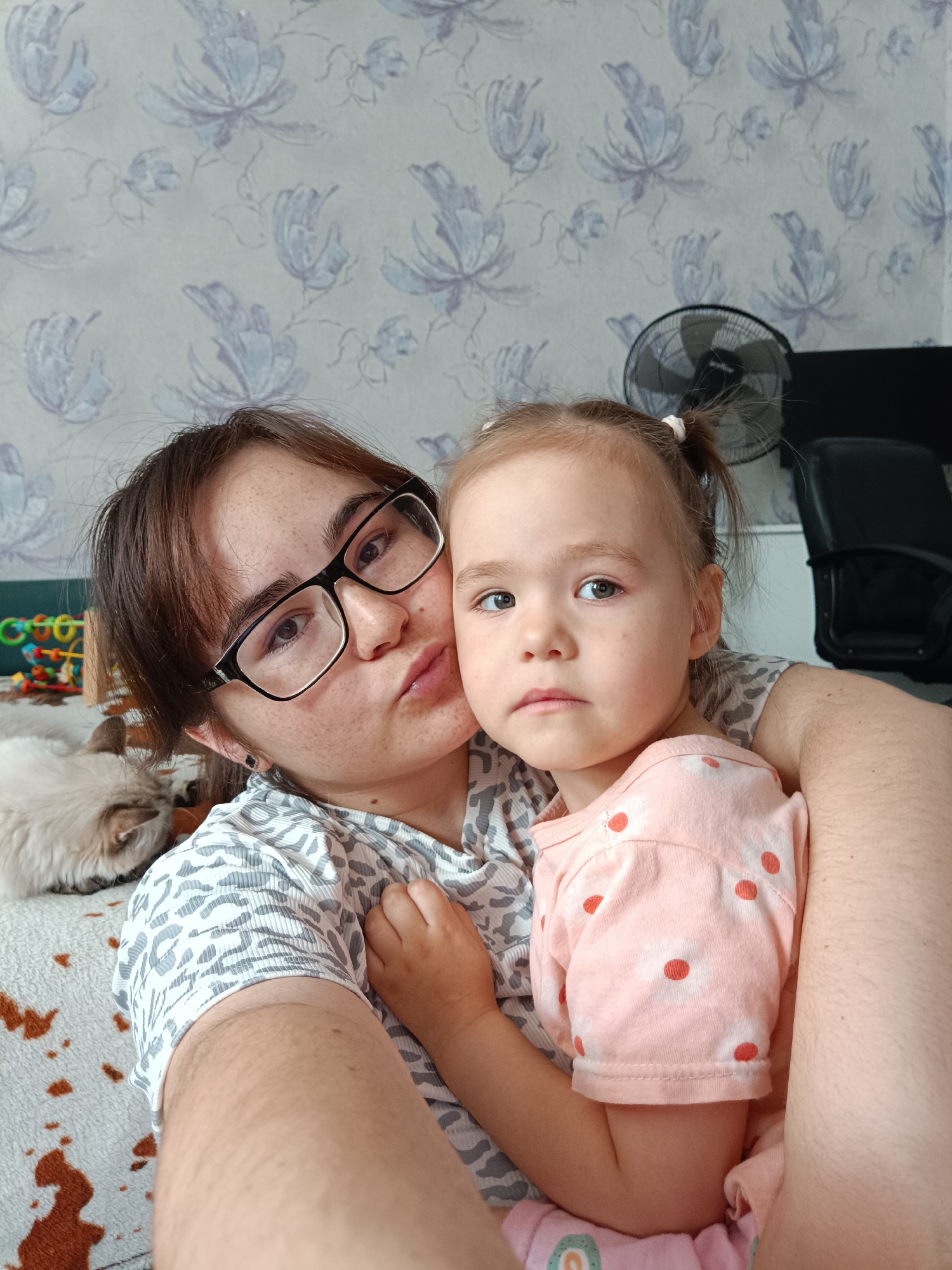 The image shows a close-up of two people, likely an adult and a child, posing for a selfie in an indoor setting. The adult on the left is wearing glasses and has brown hair. They are puckering their lips. The child on the right has blonde hair styled in two pigtails and is wearing a pink shirt with red polka dots. The background features a wall with a floral pattern and a fan. To the left, a cat is lying down on a patterned blanket. The overall scene suggests a casual, domestic environment. The location is identified as Noviy, Russia.