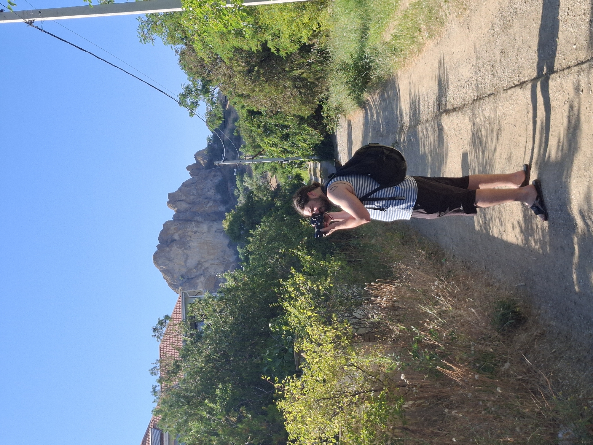 A man with a beard and a backpack is standing on a dirt road, taking a photo with a camera. He is wearing a striped tank top and shorts. The road is lined with trees and bushes, and in the background, there are rocky hills under a clear blue sky. There is a utility pole with wires on the right side of the road. A building with a red tile roof is visible behind the trees on the left. The scene appears to be in a rural or semi-rural area within the Щебетовский поселковый совет, Ukraine, given the context of the request.